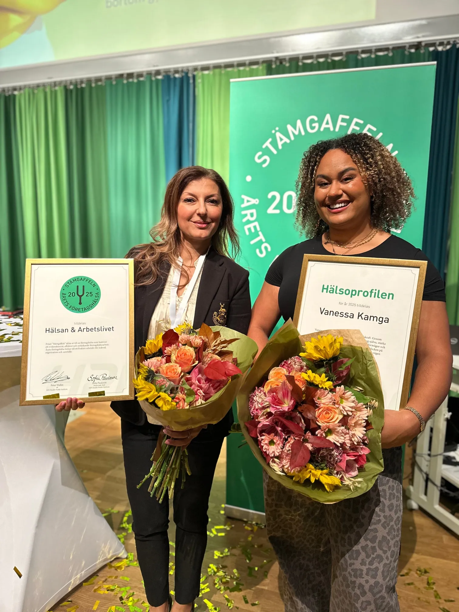 Två kvinnor står bredvid varandra med blombuketter och priscertifikat vid en prisutdelning för Hälsoprofilen 2025.