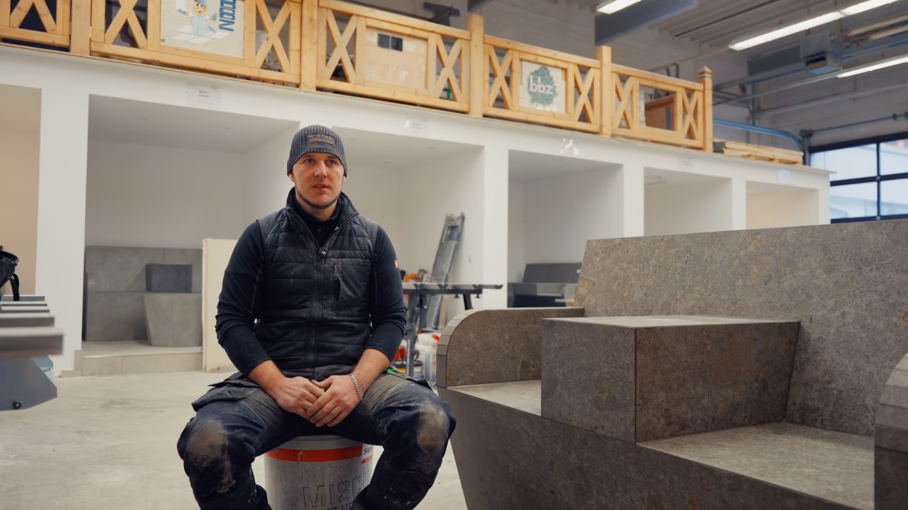 Videothumbnail mit Erfahrungsbericht von Daniel Zimon, Meisterschüler, sitzend in einer Werkstatt mit Steinarbeiten im Hintergrund.
