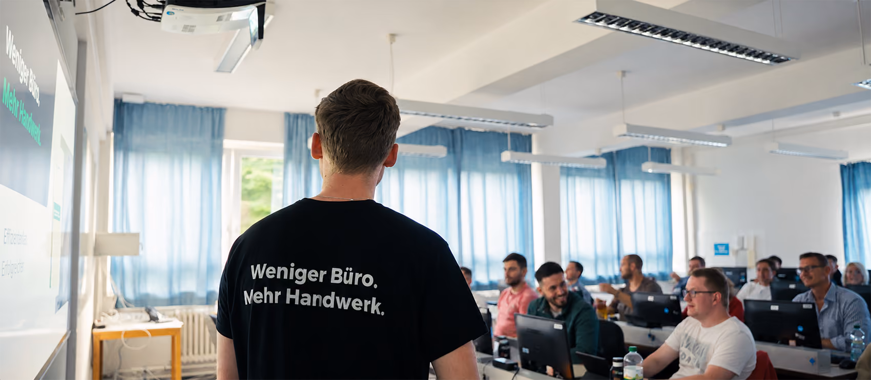Patrick Runge von plancraft steht mit dem Rücken zur Kamera vor einer Klasse und präsentiert die Software mit dem Slogan 'Weniger Büro. Mehr Handwerk.' auf seinem T-Shirt.