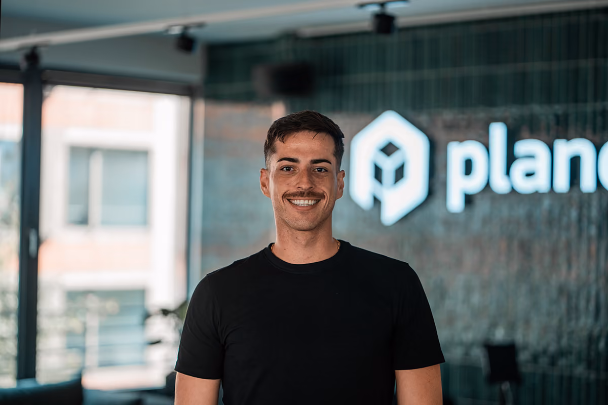 Portrait of Nick, Revenue Enablement Manager at plancraft, smiling in front of the plancraft office logo wall.