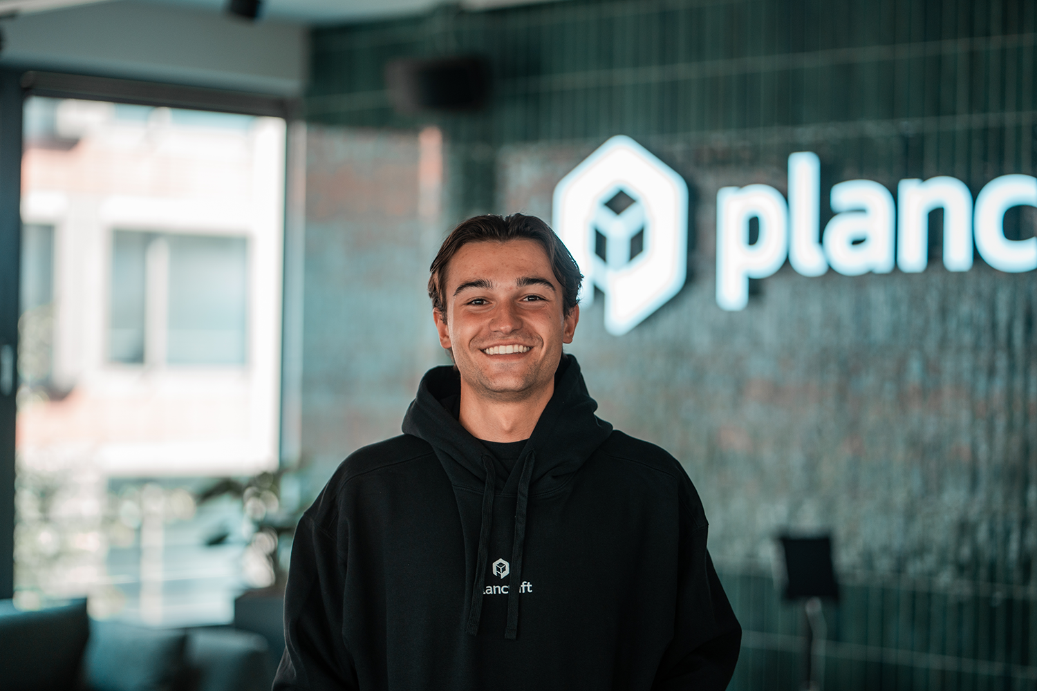 Portrait of Thiemo, SDR Netherlands at plancraft, smiling in a plancraft hoodie in front of the plancraft office logo wall.