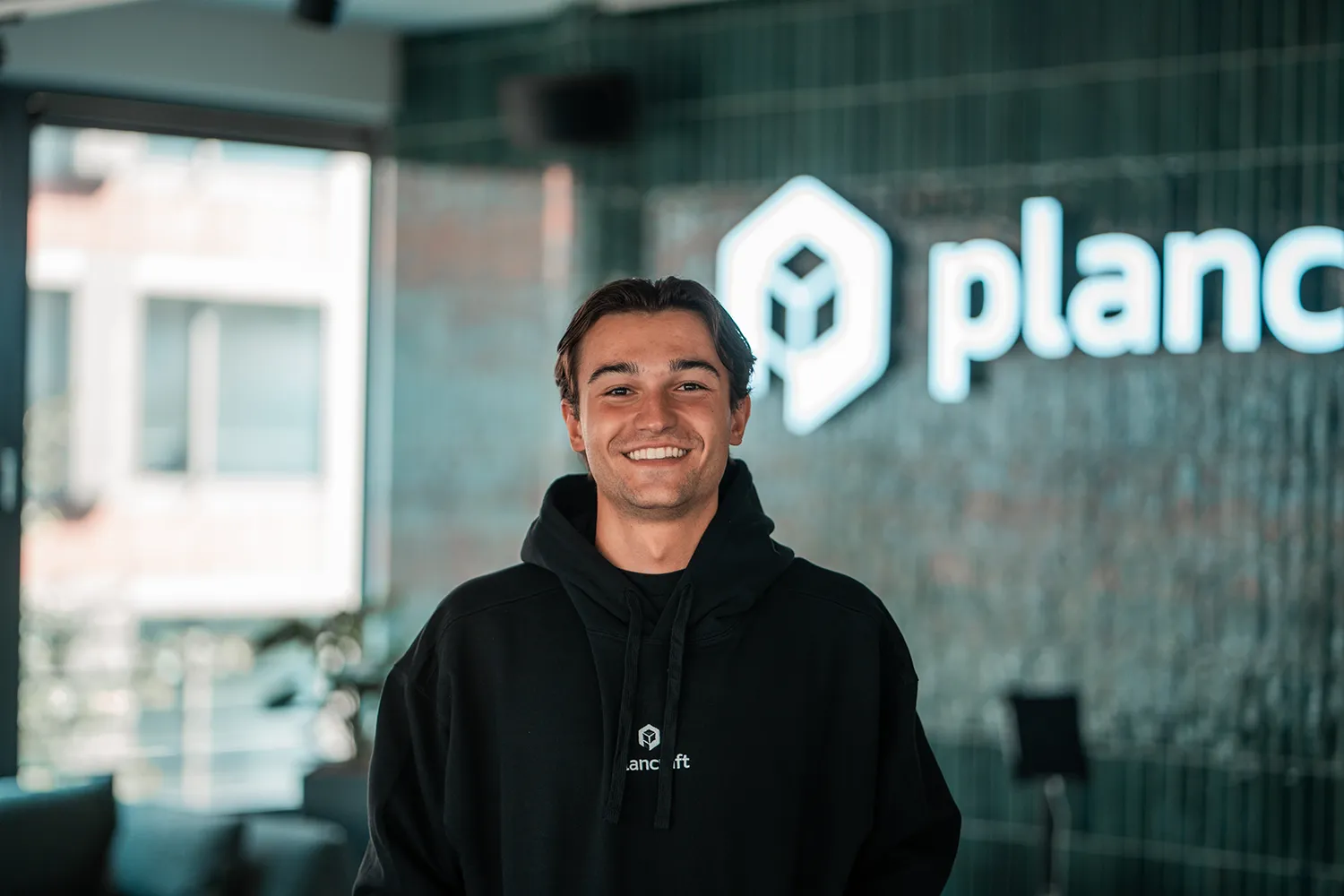 Portrait of Thiemo, SDR Netherlands at plancraft, smiling in a plancraft hoodie in front of the plancraft office logo wall.
