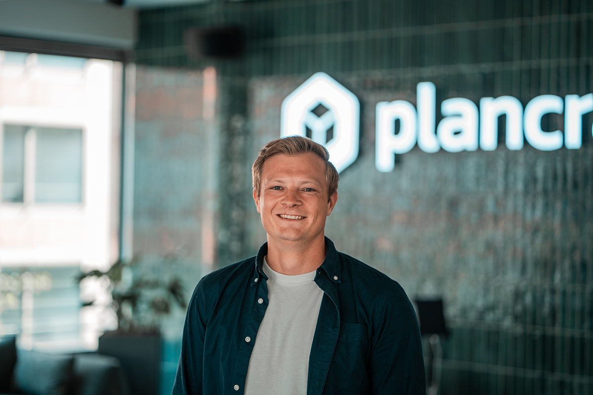 Portrait of Marvin, Engineer at plancraft, smiling in front of the plancraft office logo wall.