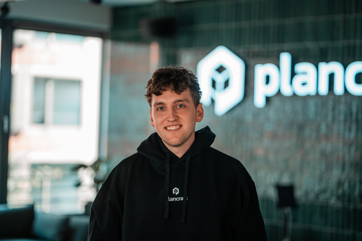 Portrait of Sebastian, SDR Vienna at plancraft, smiling in a plancraft hoodie in front of the plancraft office logo wall.