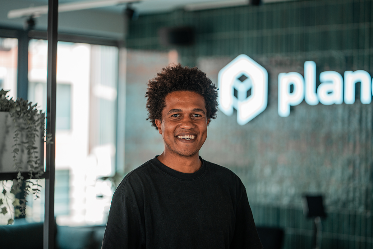 Portrait of Christian, SDR at plancraft, smiling in front of the plancraft office logo wall.