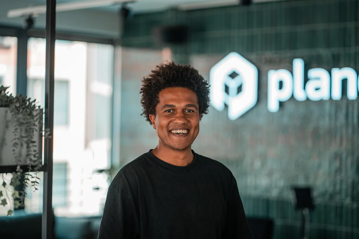 Portrait of Christian, SDR at plancraft, smiling in front of the plancraft office logo wall.