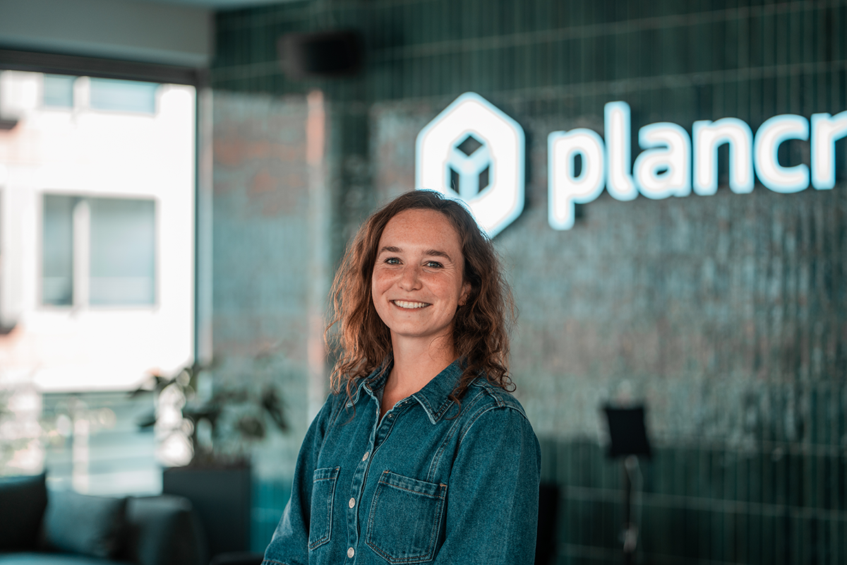 Portrait of Sandra, SDR at plancraft, smiling in a denim shirt in front of the plancraft office logo wall.
