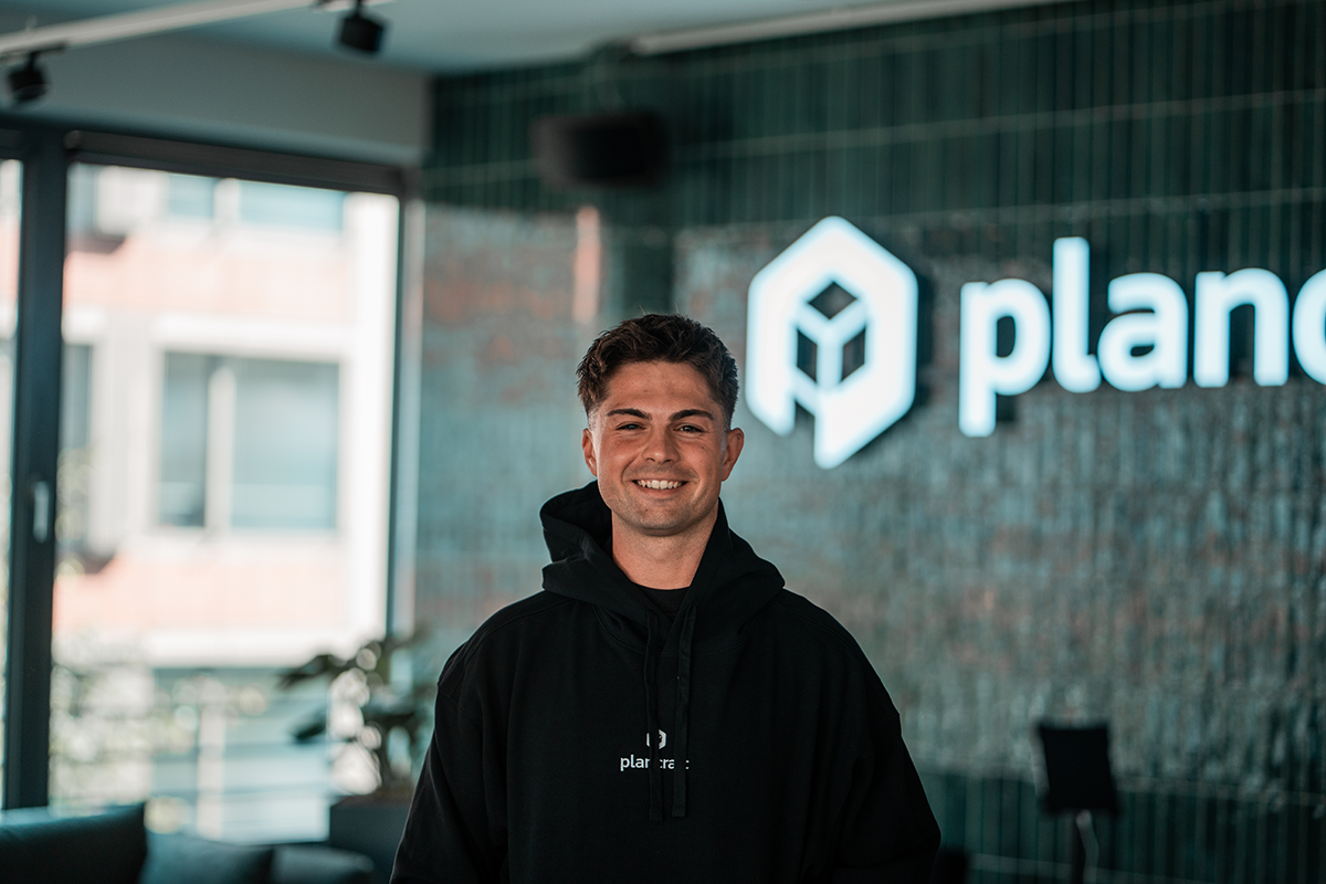 Portrait of Matteo, SDR at plancraft, smiling in a plancraft hoodie in front of the plancraft office logo wall.