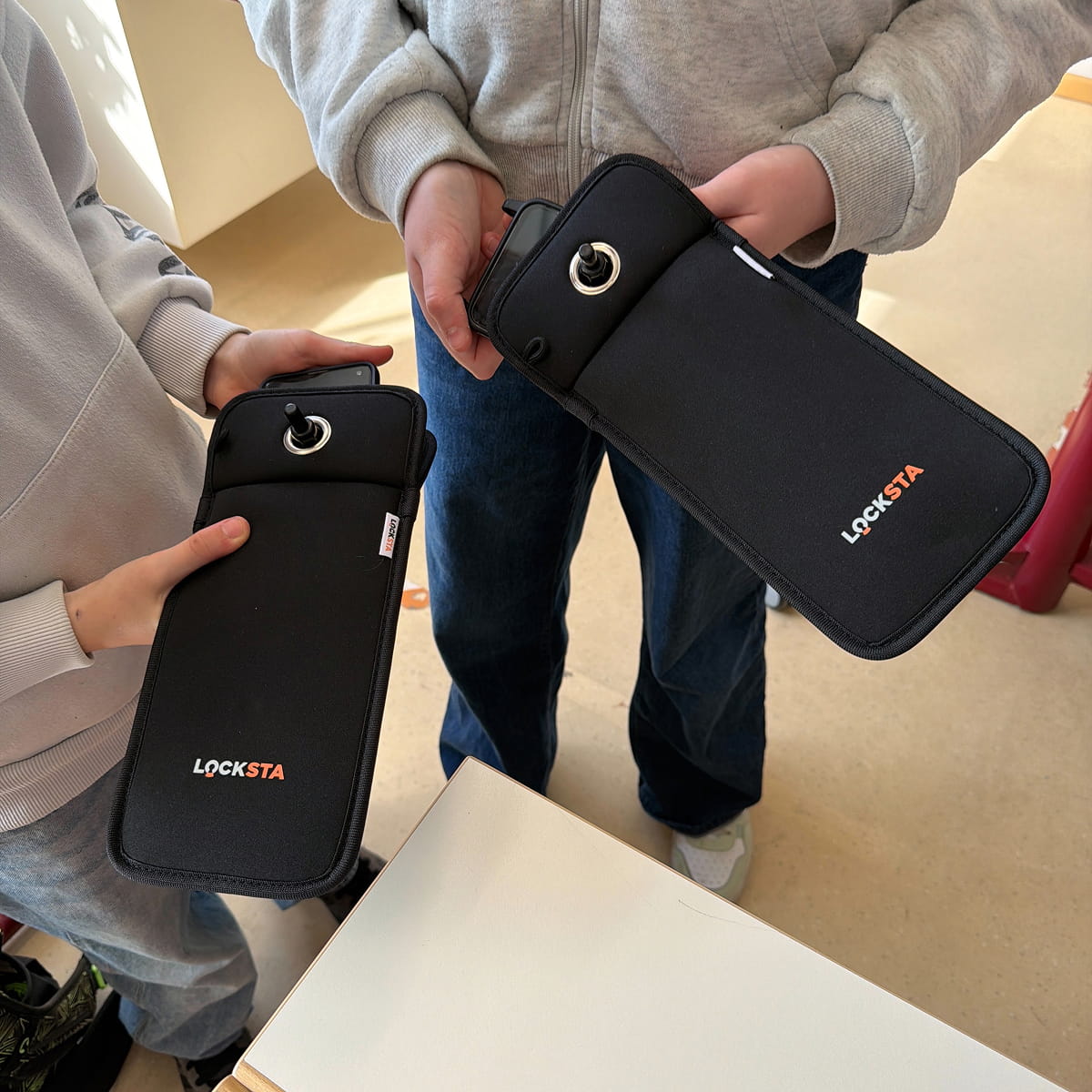Zwei Personen halten jeweils eine schwarze Locksta-Handytasche mit einem Drehschalter, eine Person legt ein Smartphone in die Tasche.