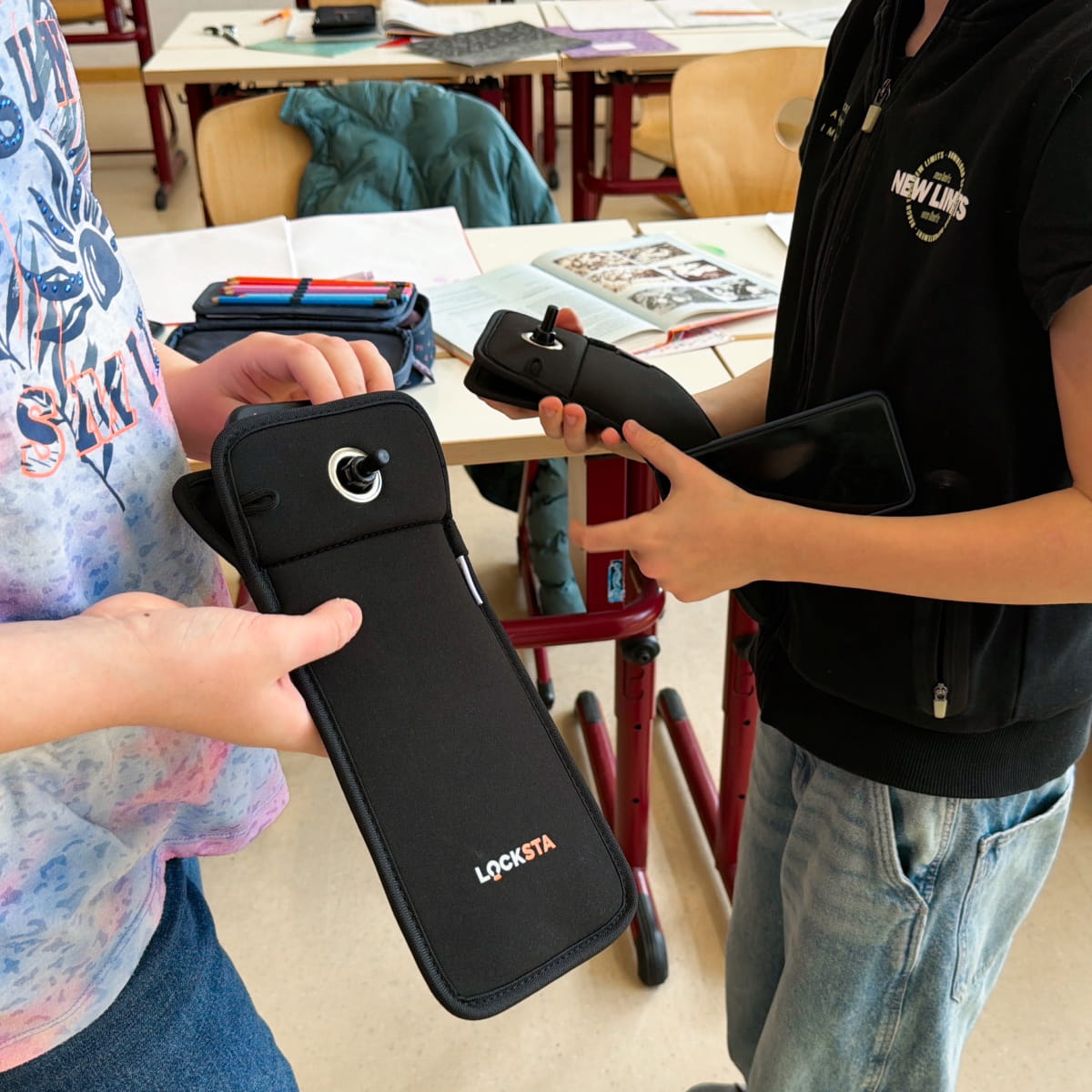 Zwei Kinder in einem Klassenzimmer halten schwarze LOCKSTA-Handytaschen, eine mit einem Smartphone darin.