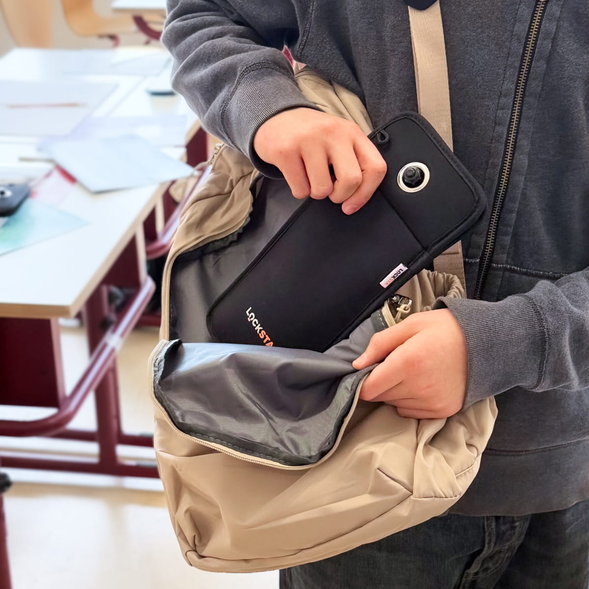 Person steckt eine schwarze Locksta-Handytasche in eine beige Umhängetasche vor einem Klassenzimmer.