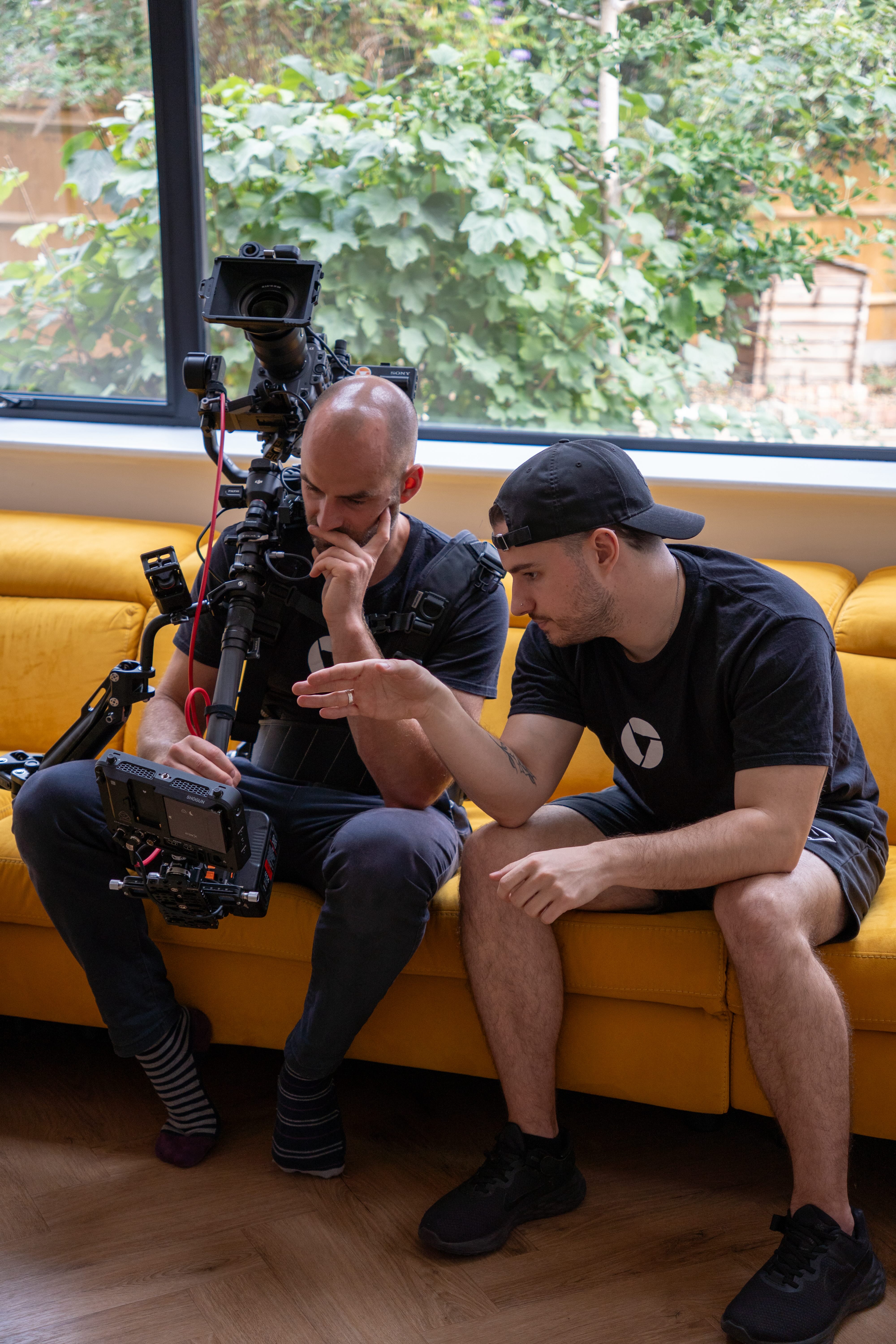 Two men sitting on a yellow couch reviewing footage on a professional video camera rig, with a green garden visible through a window behind them.