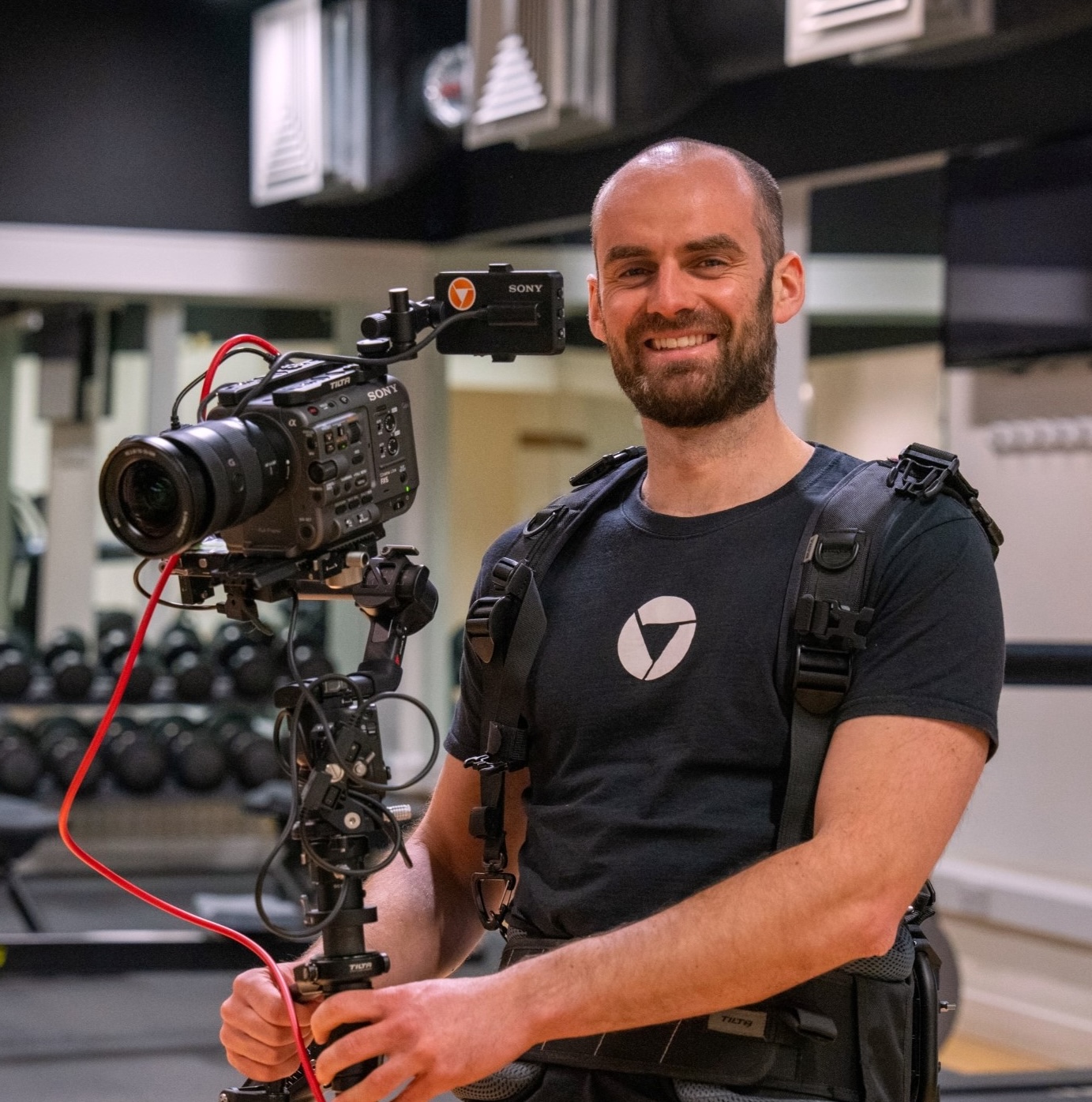 Smiling man wearing a stabilization harness holding a professional Sony video camera in a gym setting.