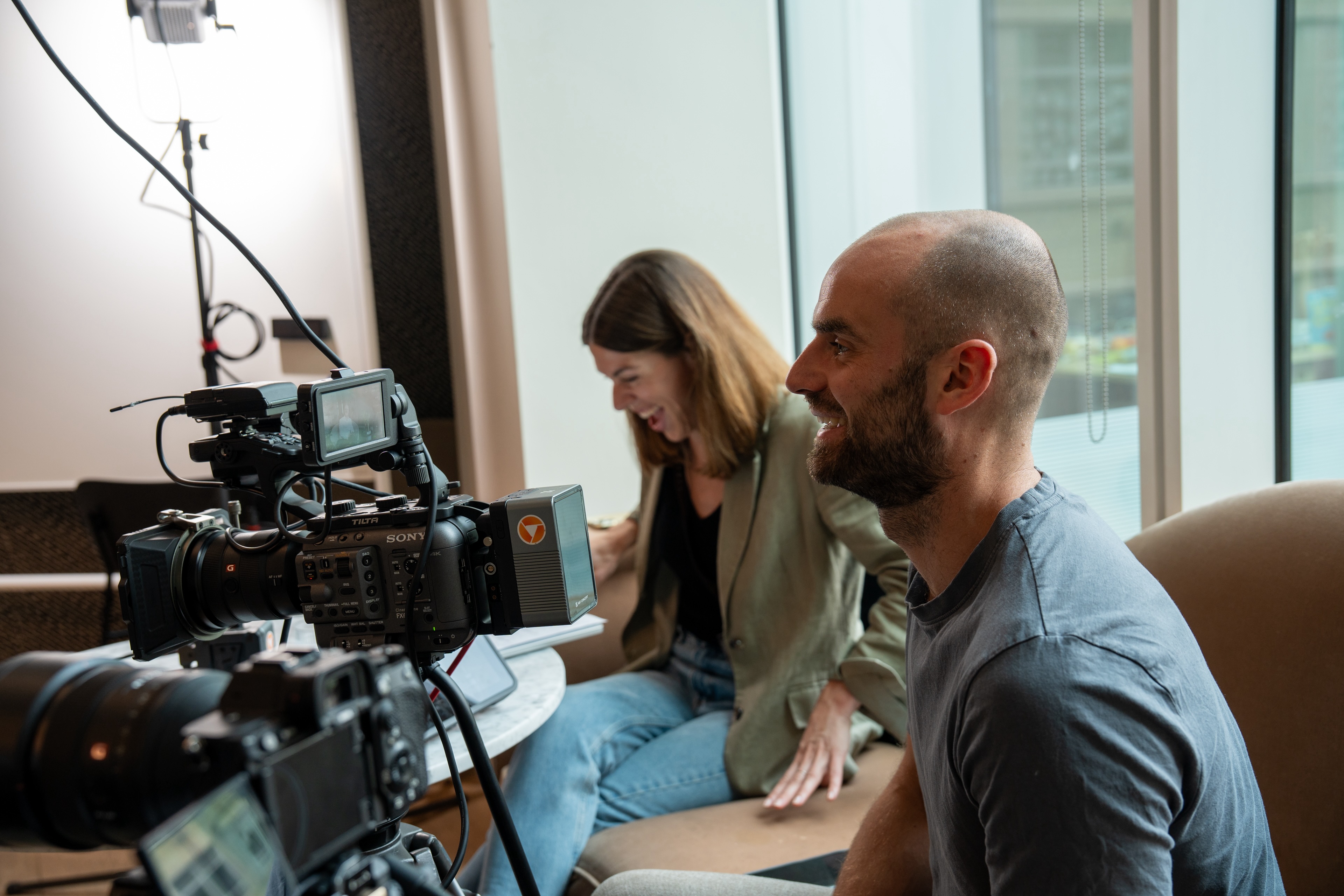 Man and woman sitting indoors near cameras, the woman laughing and the man smiling.