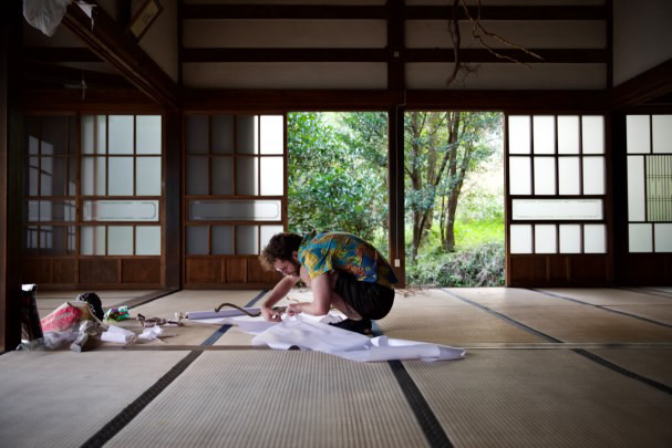 artist working on the floor of a japanese temple surrounded by art supplies. behind him the door opens onto a view of forest