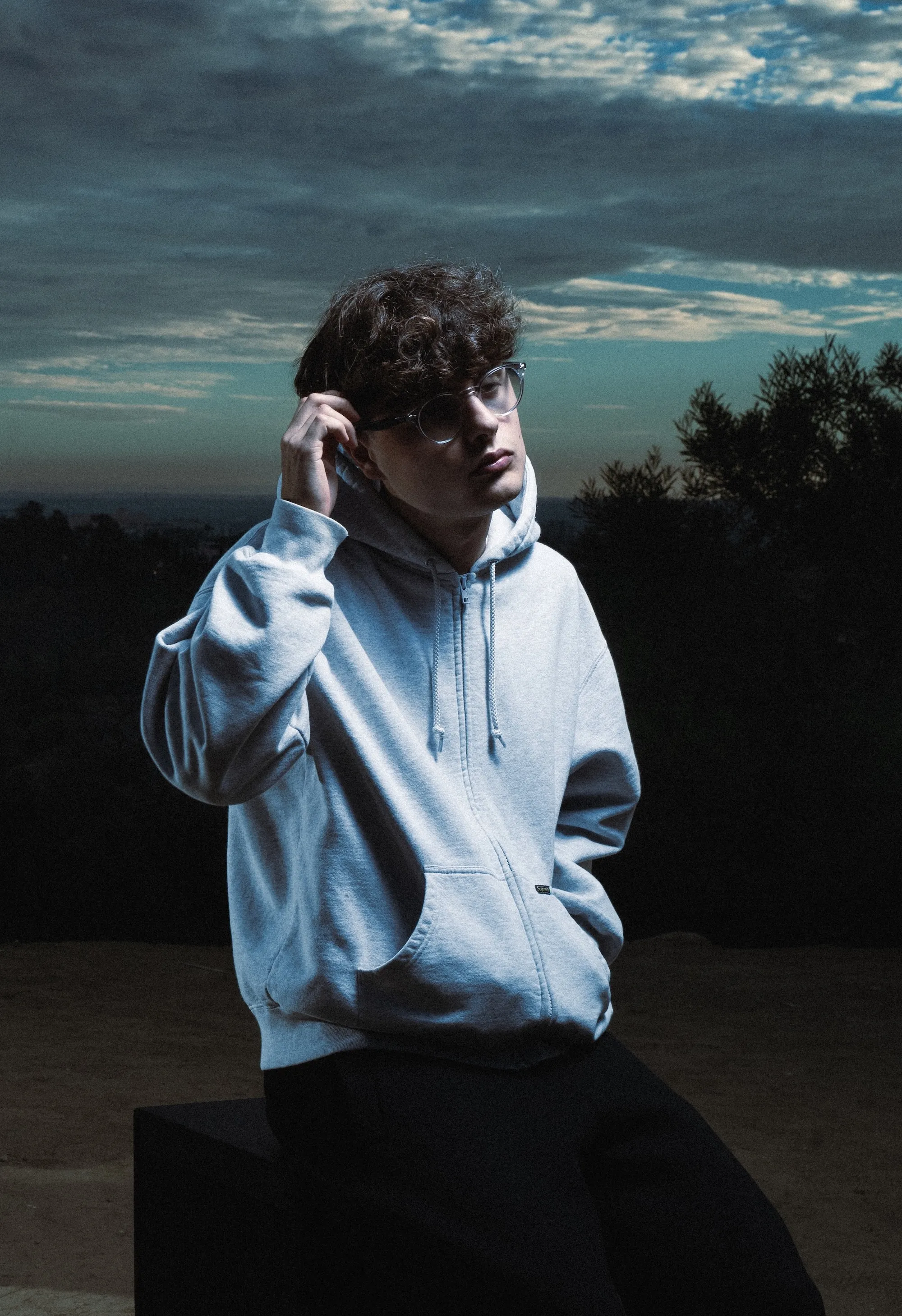 Person wearing light blue hoodie and sunglasses against dramatic cloudy sky and trees