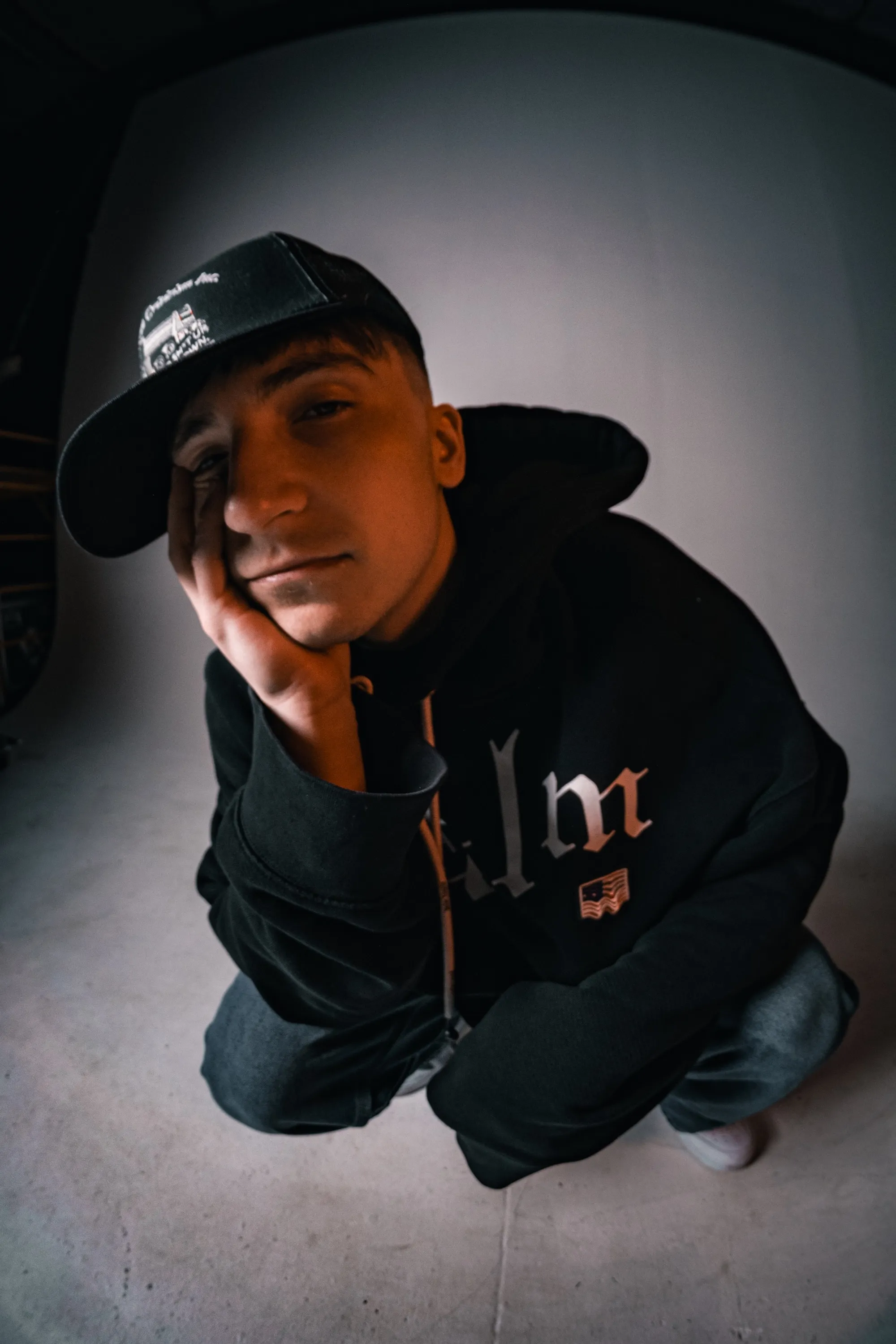 Young man wearing black cap and hoodie crouching, smiling at camera from above