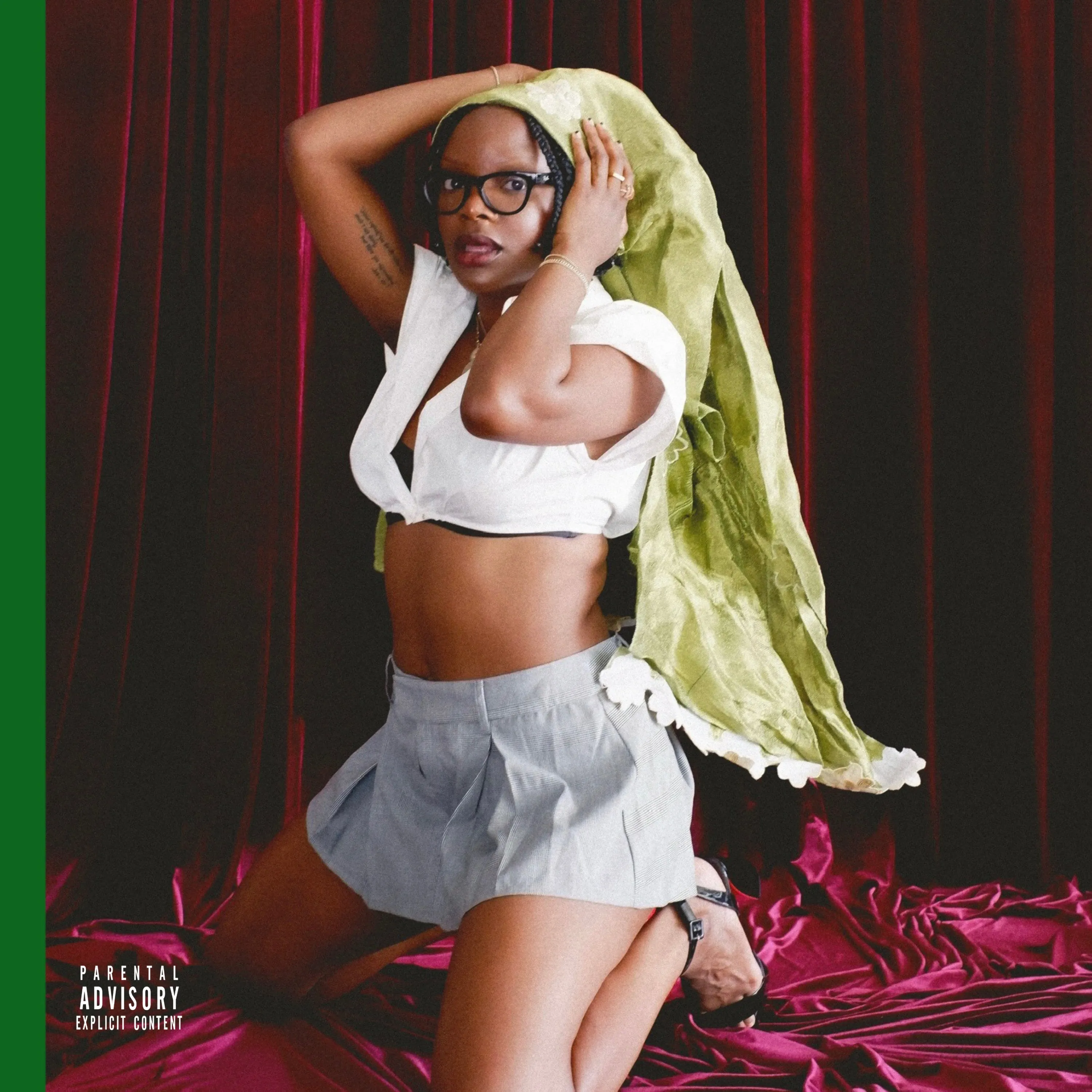 Person wearing glasses and white crop top poses with green fabric against red velvet curtains