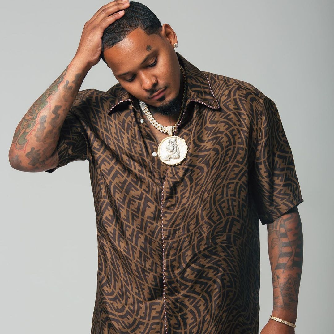 Man with tattoos wearing patterned shirt and pendant necklace, hand on head