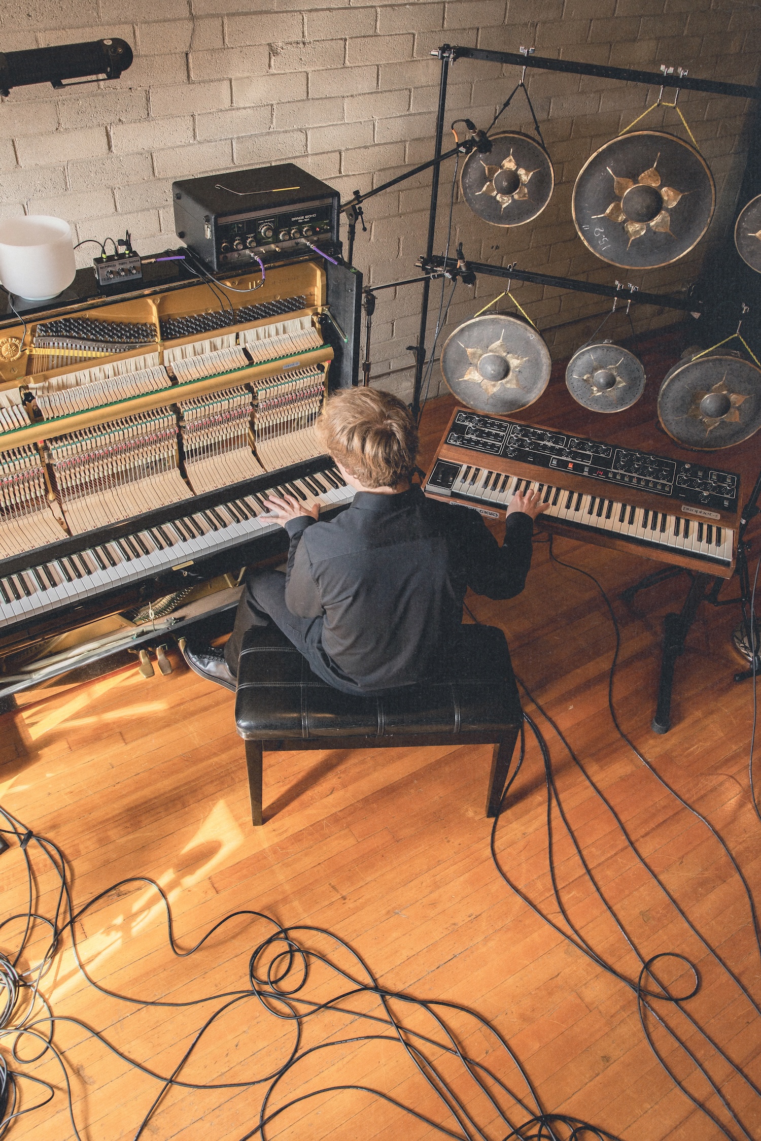 Musician seated at piano in studio with keyboards, synthesizers, and hanging drum pads