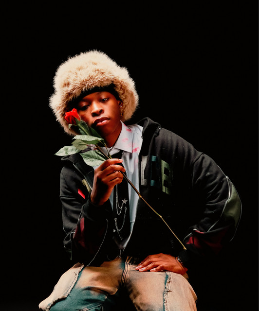 Young person wearing cream fluffy hat and black jacket, holding red rose against black background