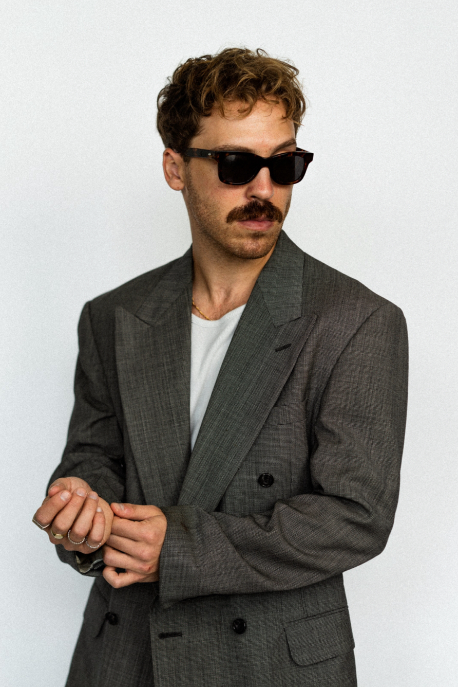 Man wearing dark sunglasses, gray blazer, and rings posing against white background.