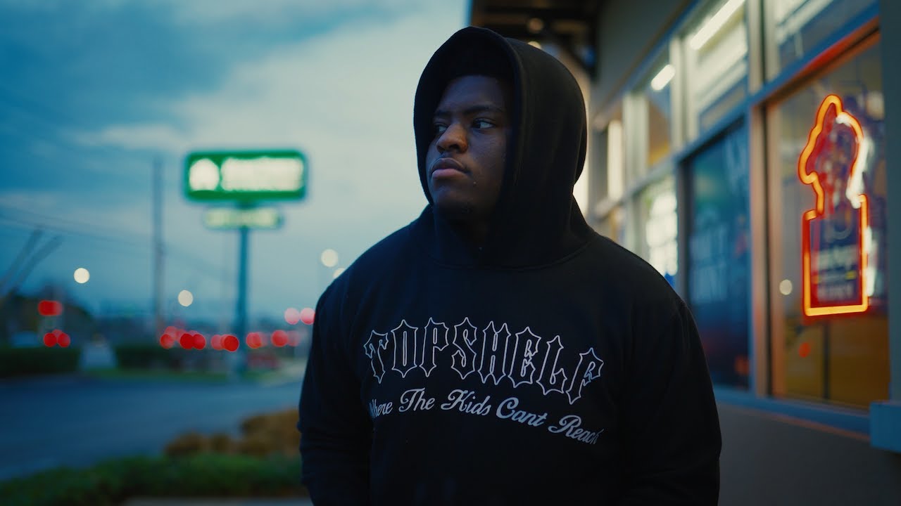 Man in dark hoodie stands on urban street at dusk with neon signs