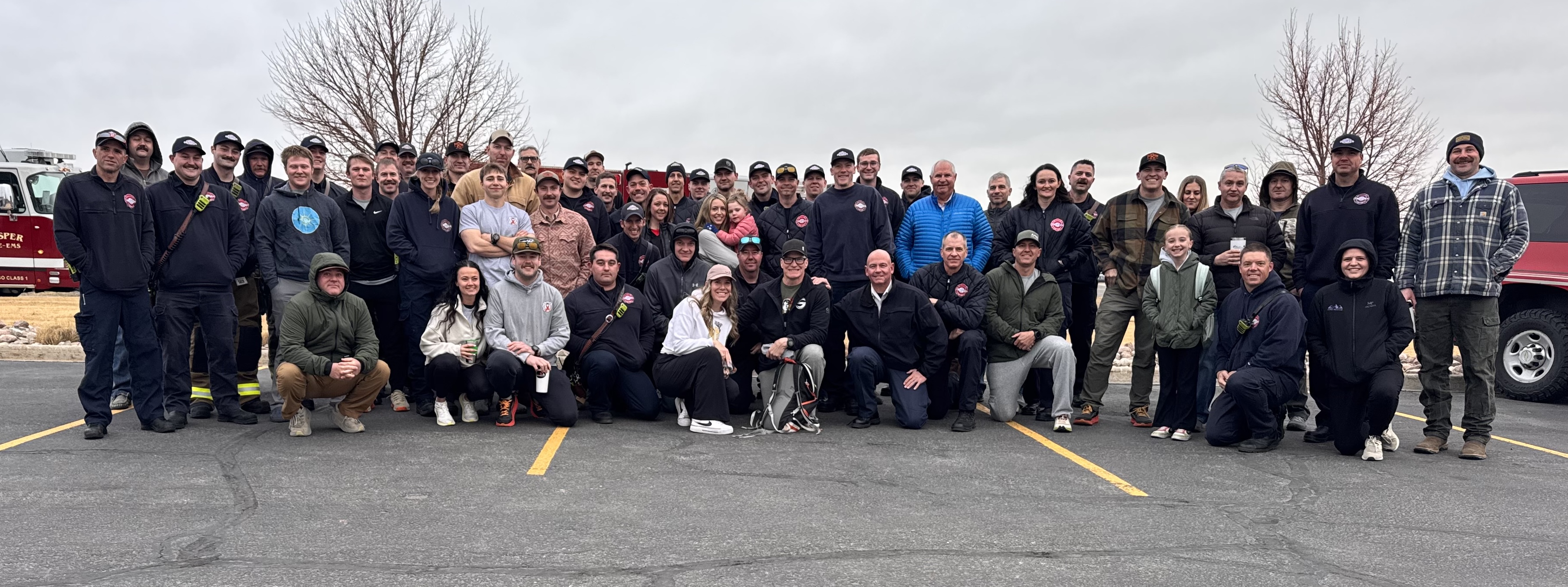 Large group of people posing together outdoors on a cloudy day in a parking lot with two leafless trees and vehicles in the background.