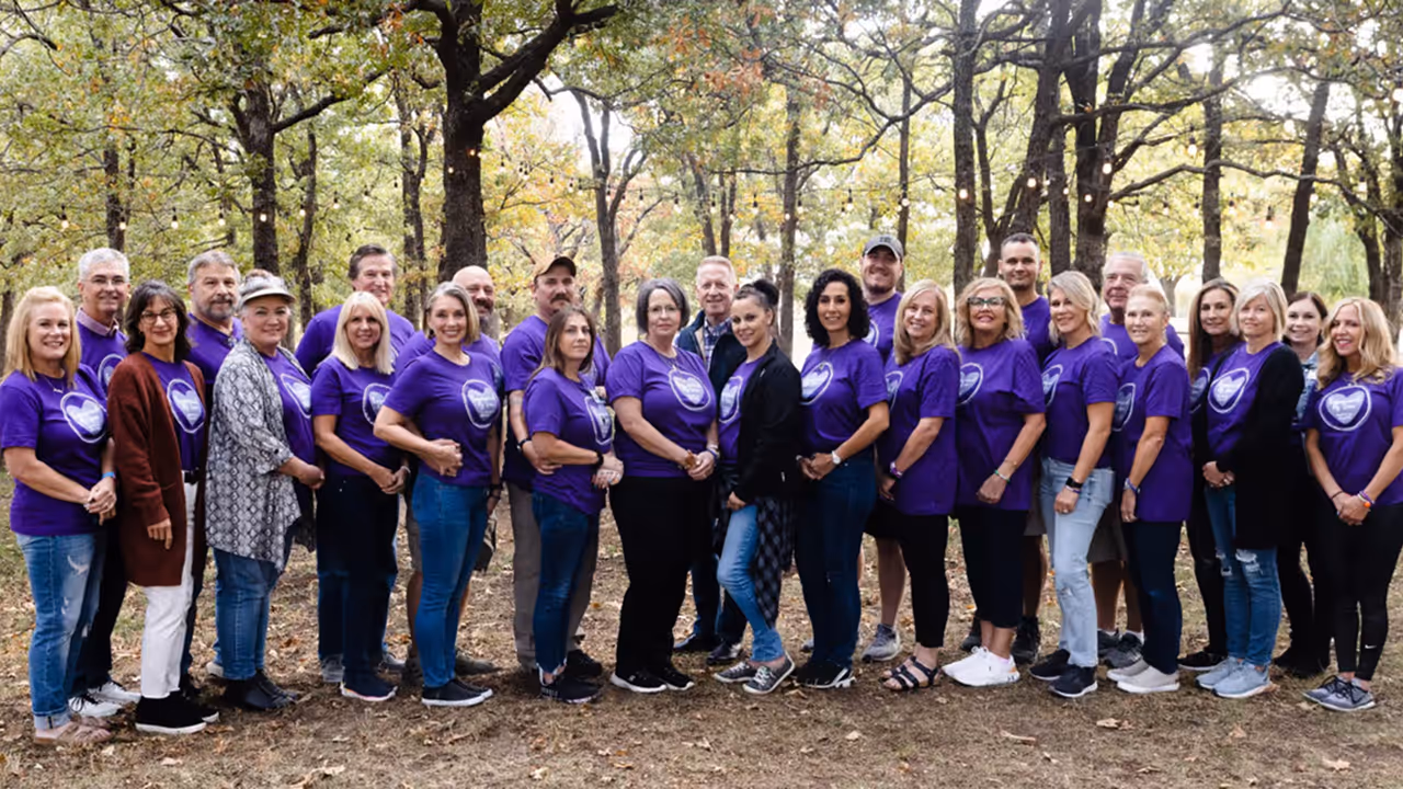 Group of adults standing outdoors in a wooded area, wearing purple Hope After Loss t-shirts with heart logos, smiling at the camera.