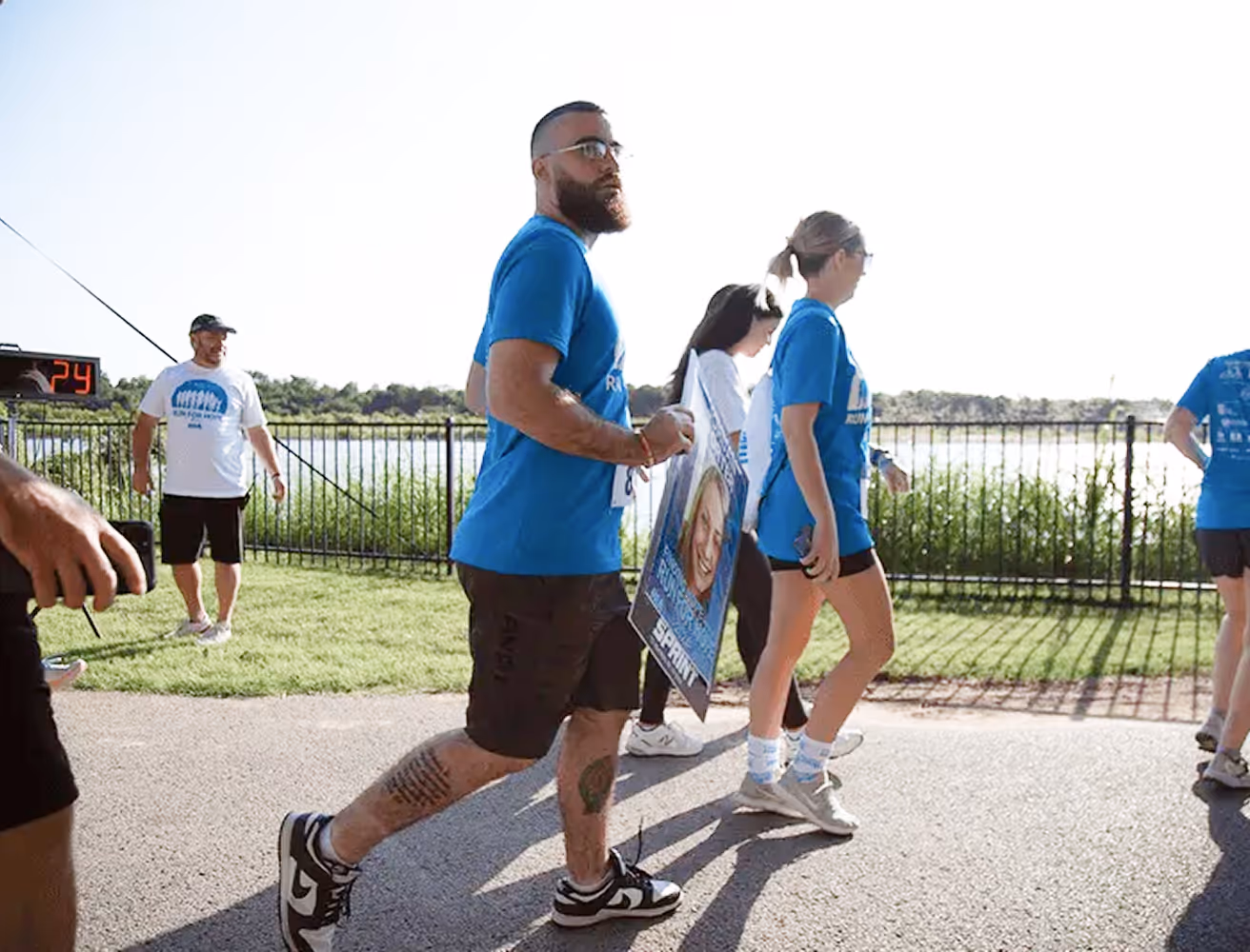 Group of people walking outdoors near a lake during the run for hope, one man in a blue shirt holding a poster with a woman's photo.