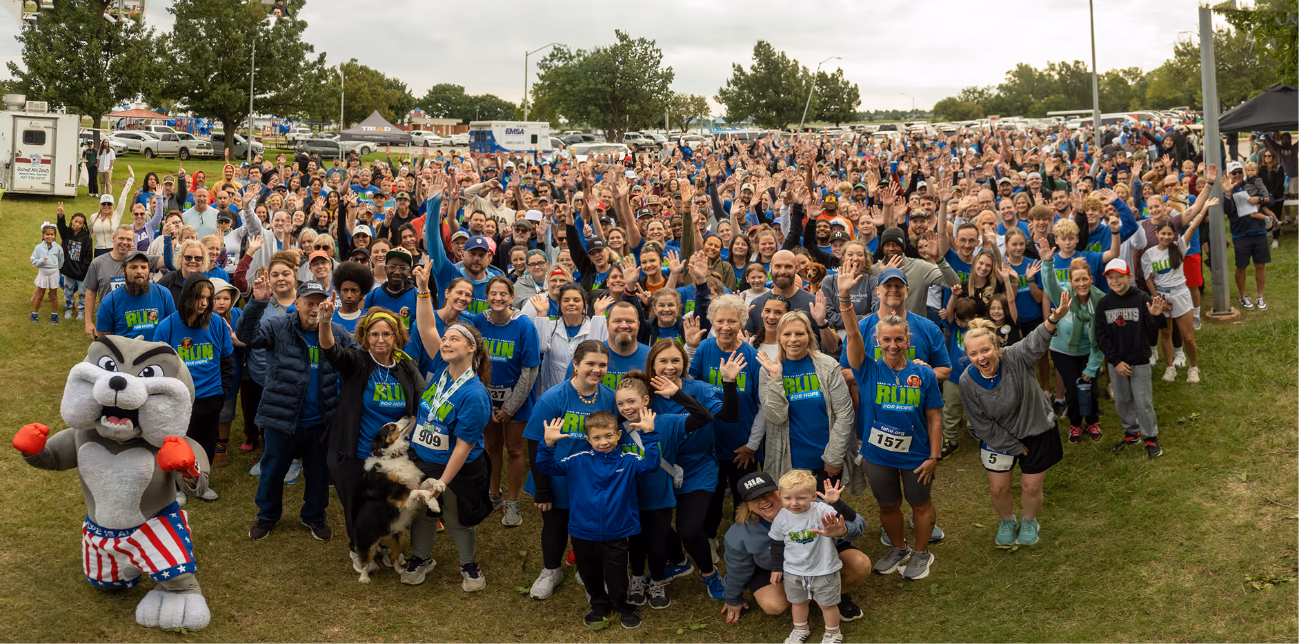 Large group of people outdoors waving at the camera, many wearing blue 'Run for Hope' shirts, with a bulldog mascot in red boxing gloves and patriotic shorts in front.