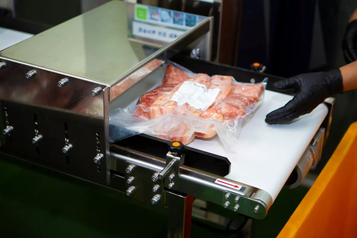 진공포장된 고기 팩이 컨베이어 벨트 위에 놓여 있고 검은 장갑을 낀 손이 근처에 위치해 있음.
