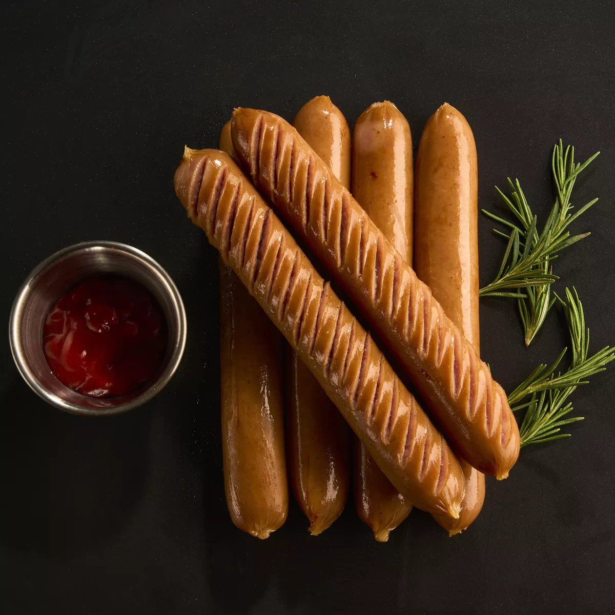 검은 배경 위에 잘게 칼집 낸 소시지 다섯 개와 케첩이 담긴 금속 컵, 심플한 로즈마리 가지 두 개가 놓여 있음.