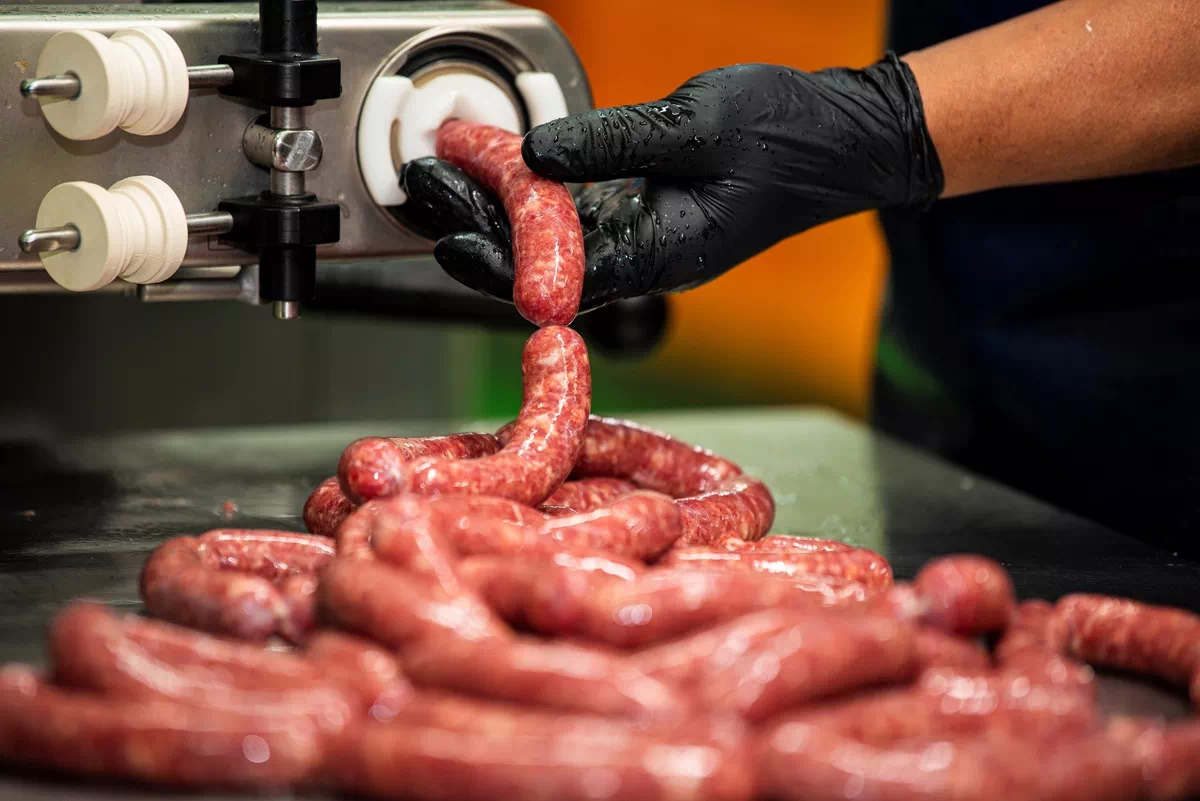 검은 장갑을 낀 손이 소시지 충전 기계에서 갓 만들어진 소시지를 잡고 있는 모습.