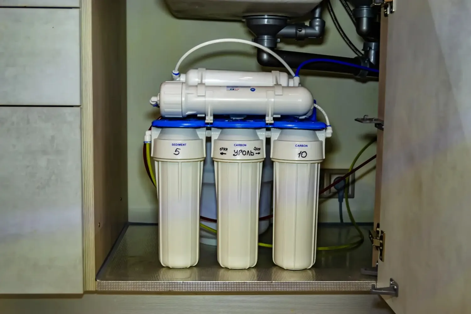 A multi-stage under-sink water filtration system with three tall white filter cartridges labeled "SEDIMENT," "CARBON," and a third with unreadable text, is installed beneath a kitchen sink.