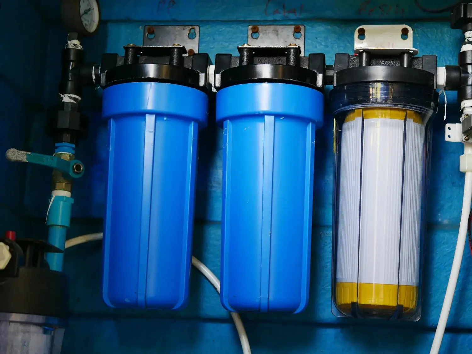  Three large water filter housings, two blue opaque and one clear with a yellow filter cartridge inside, are mounted on a blue wall.
