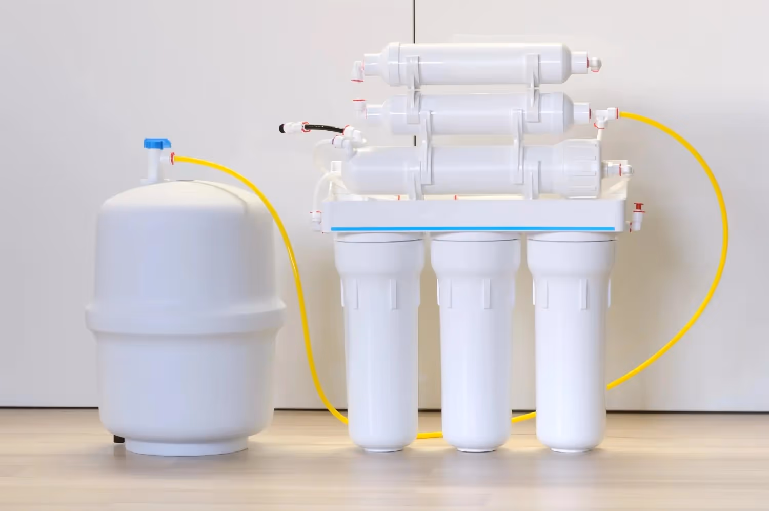 A complete home water purification system, including a large white tank and a multi-stage filter unit with white cartridges and yellow tubing, sits against a white wall.