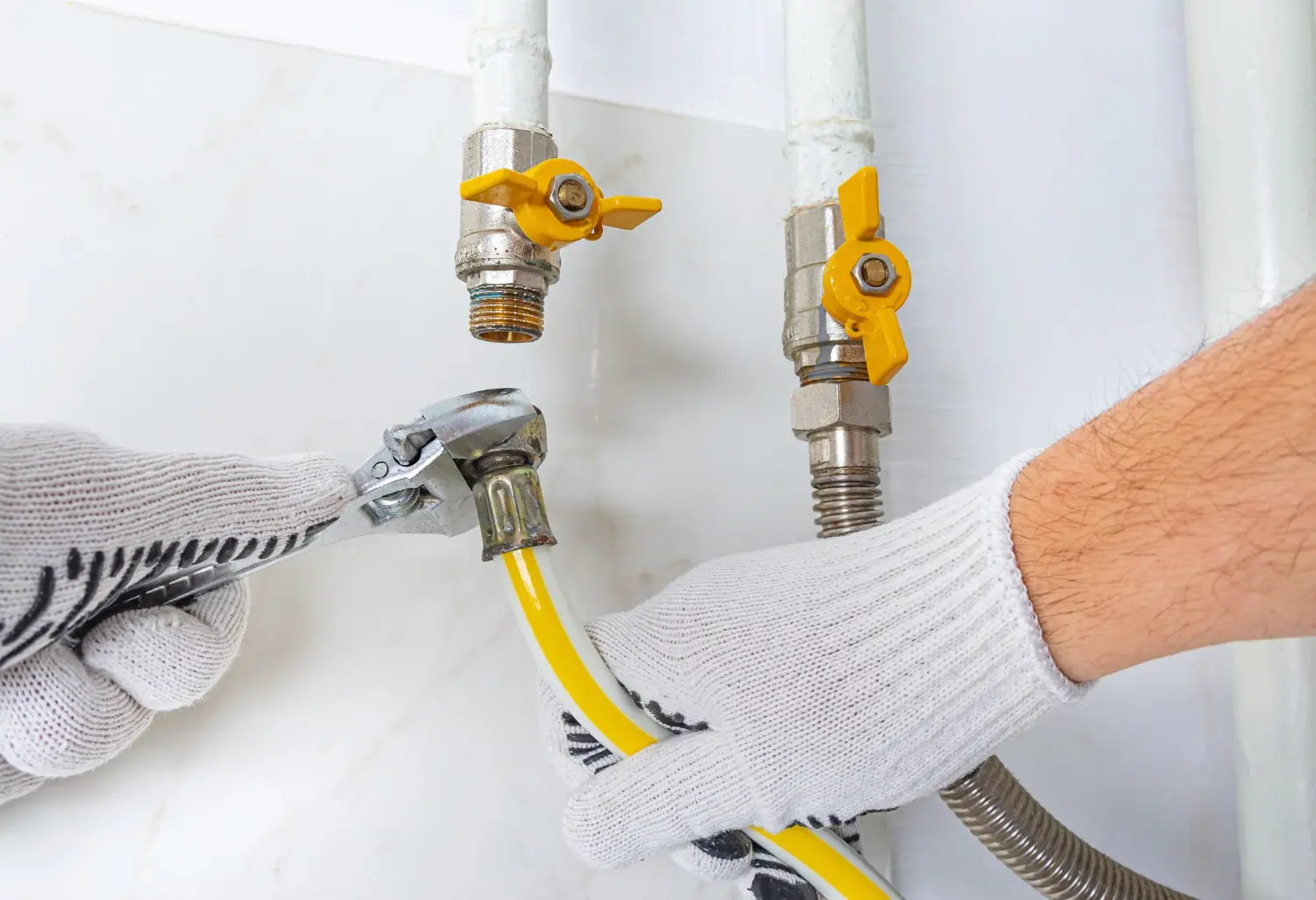 A person wearing white gloves is using a wrench to connect a yellow flexible gas line to a white gas pipe with a yellow shut-off valve.