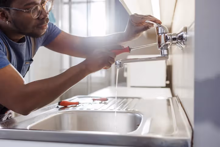 A person wearing glasses is using a screwdriver to fix a leaky faucet over a stainless steel sink, with water dripping from the tap.