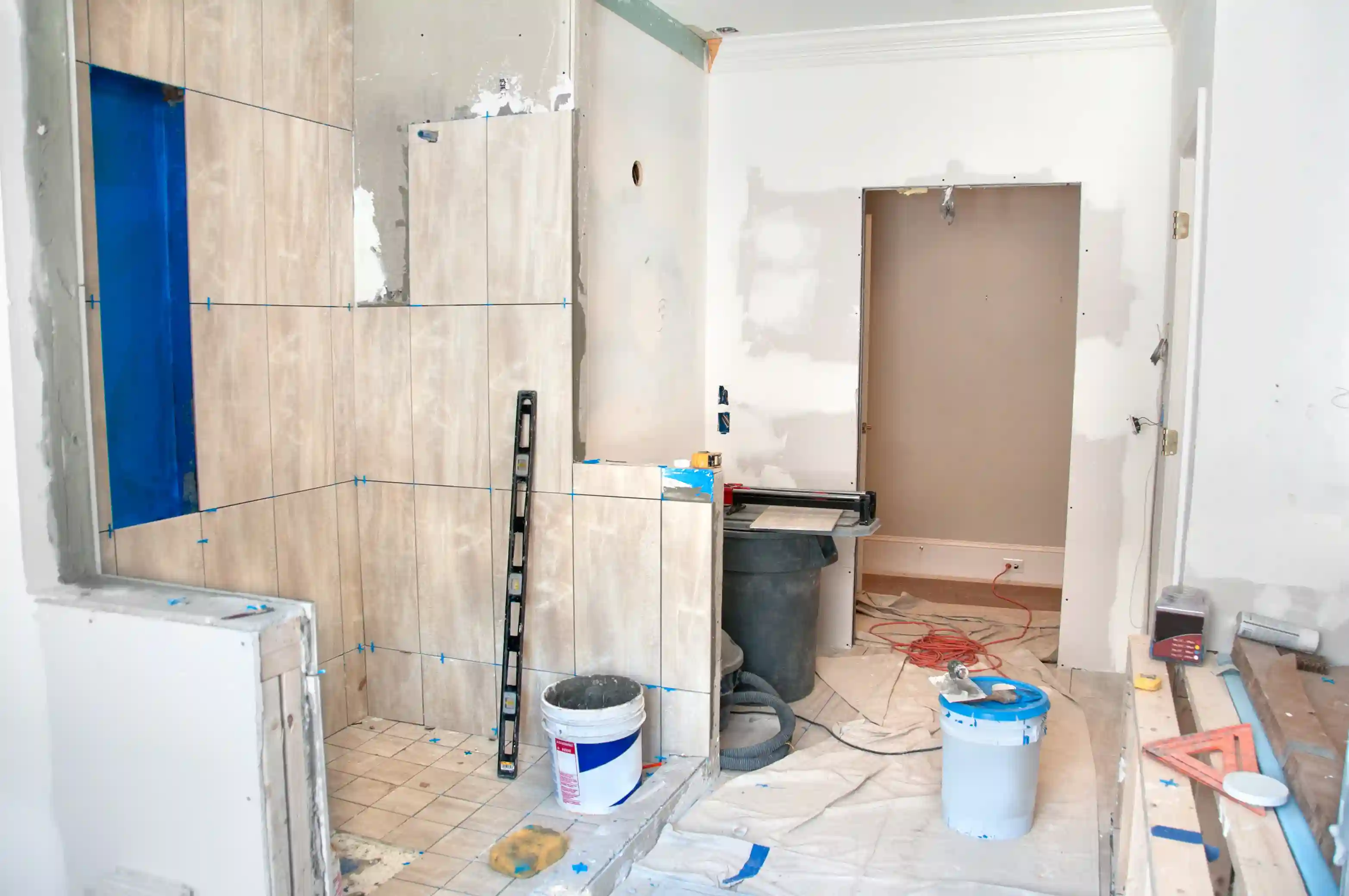 Bathroom renovation in progress with wall tiles, blue spacers, level tool, and construction materials on protective floor sheet
