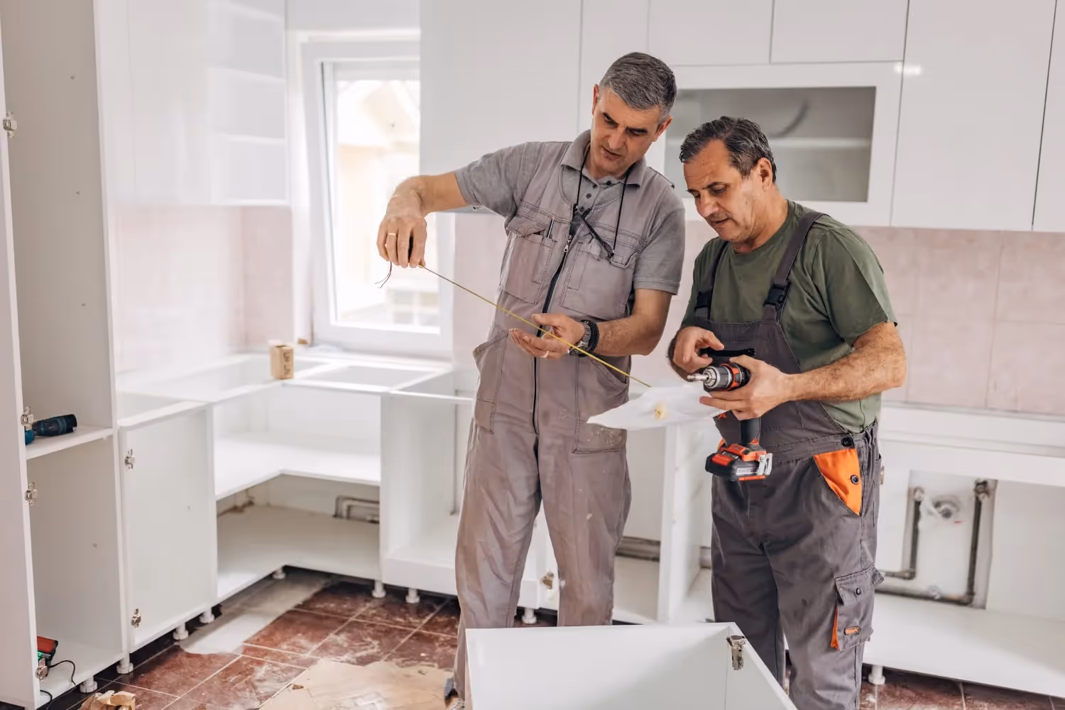 Two male construction workers are standing in a kitchen under renovation, assembling white cabinets, with one holding a drill and the other a tape measure.
