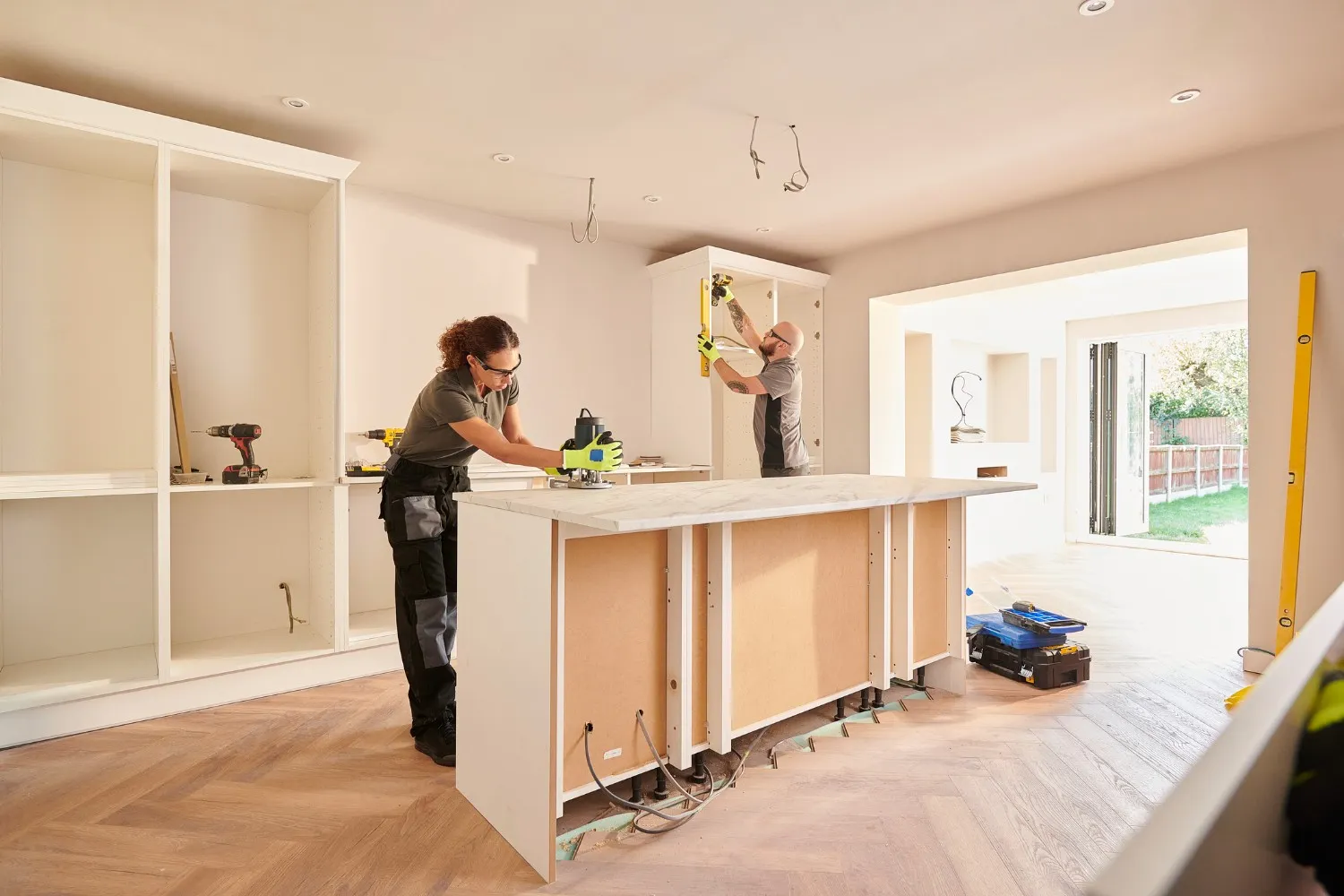 Two workers are in a kitchen under renovation; a woman is using an electric planer on a countertop, while a man in the background works on upper cabinets.