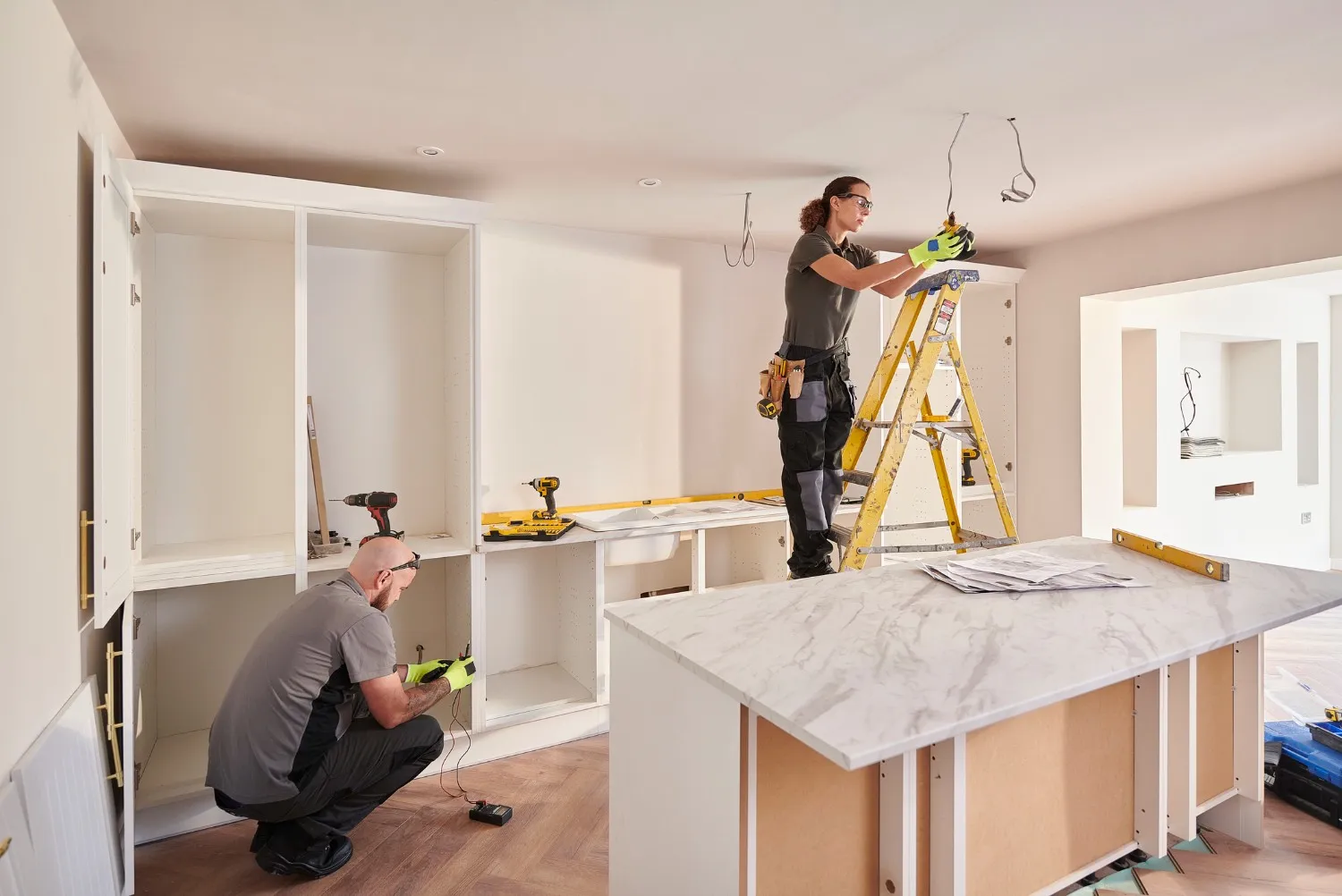 Two workers are in a kitchen under renovation; a man is kneeling and connecting wires in a lower cabinet, while a woman on a yellow ladder works on electrical wiring in the ceiling.
