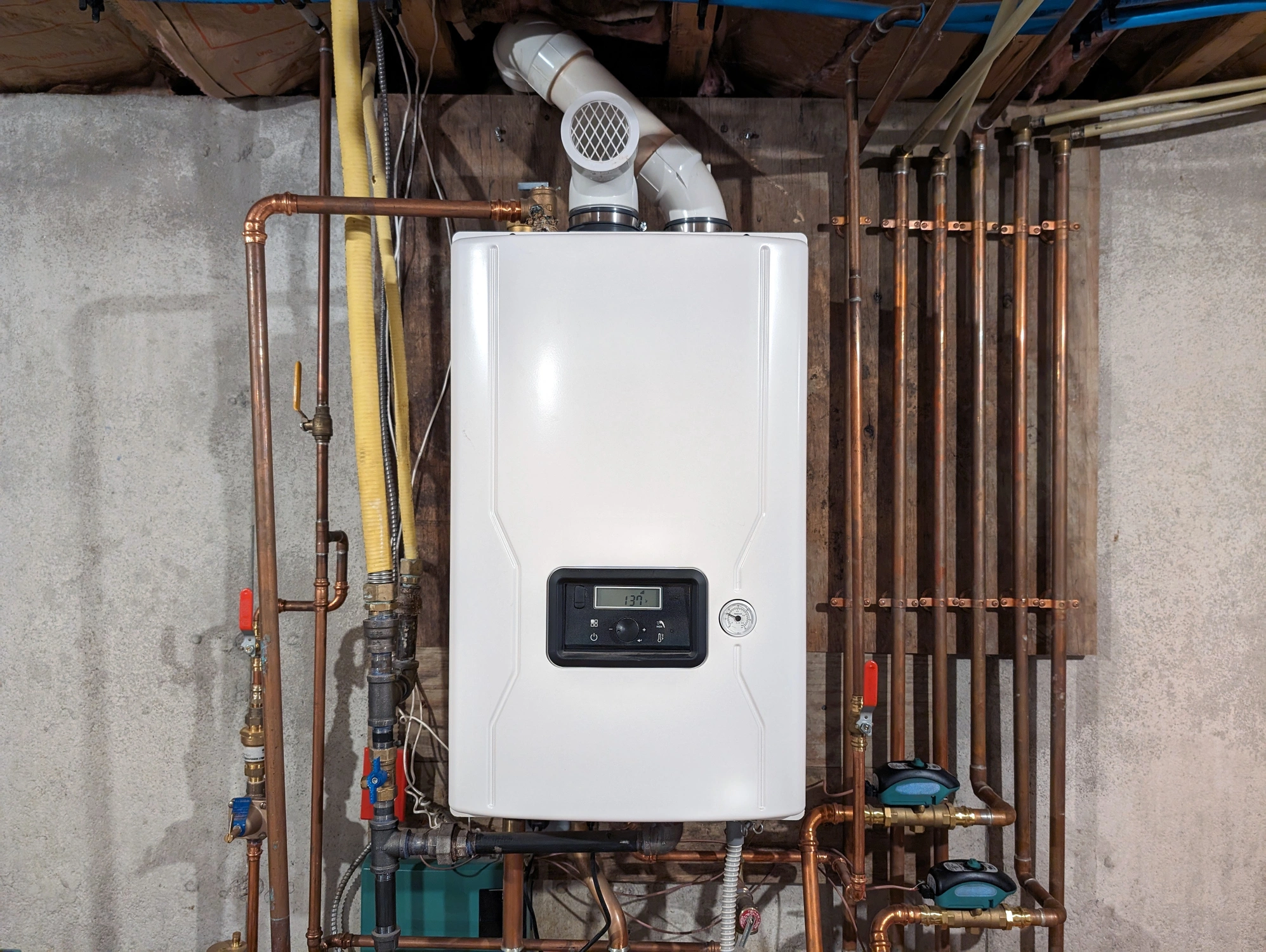 A white tankless water heater is mounted on a concrete wall, surrounded by copper pipes.
