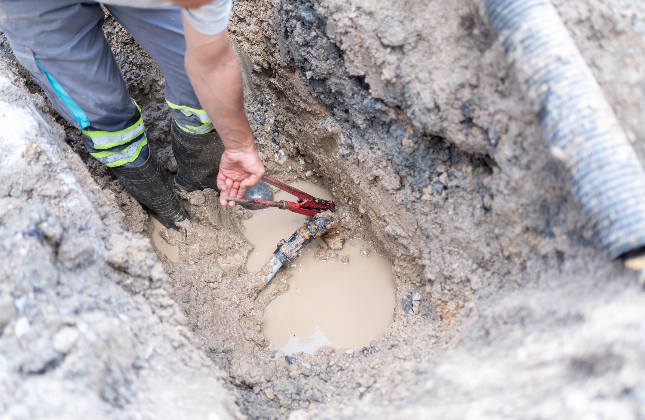 Worker in reflective gear uses a red pipe wrench to repair a leaking water pipe in a muddy trench.