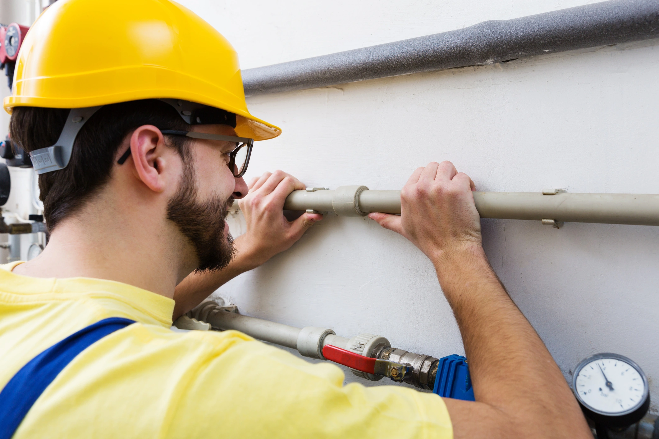 A worker in a yellow hard hat and protective goggles examines a pipe on a wall