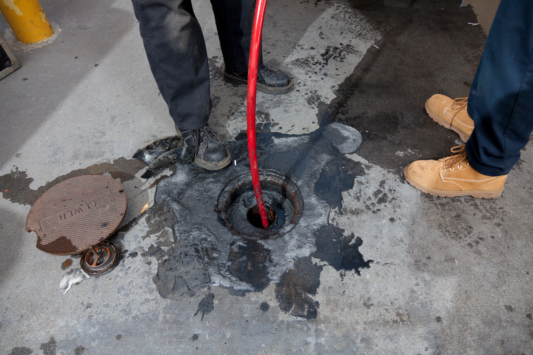 Two people stand by an open manhole. One person uses a red cable, likely for drainage work. Dark stains surround the area, indicating a spill.
