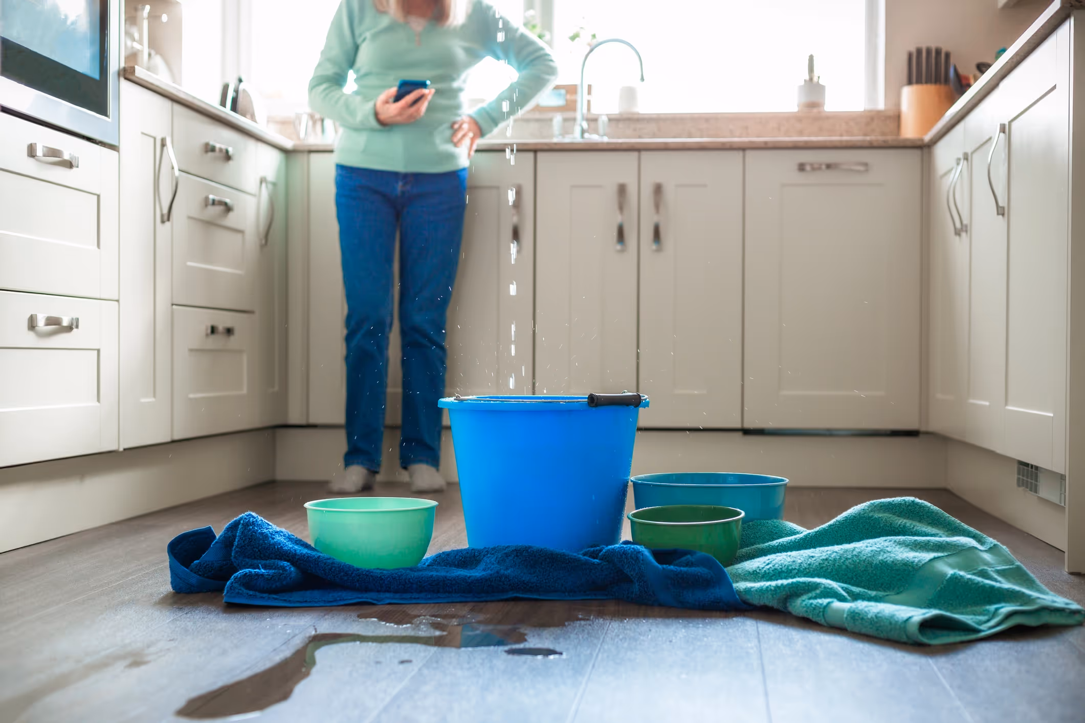 A woman stands in a kitchen looking at her phone, concerned. Water drips from the ceiling into a blue bucket,
