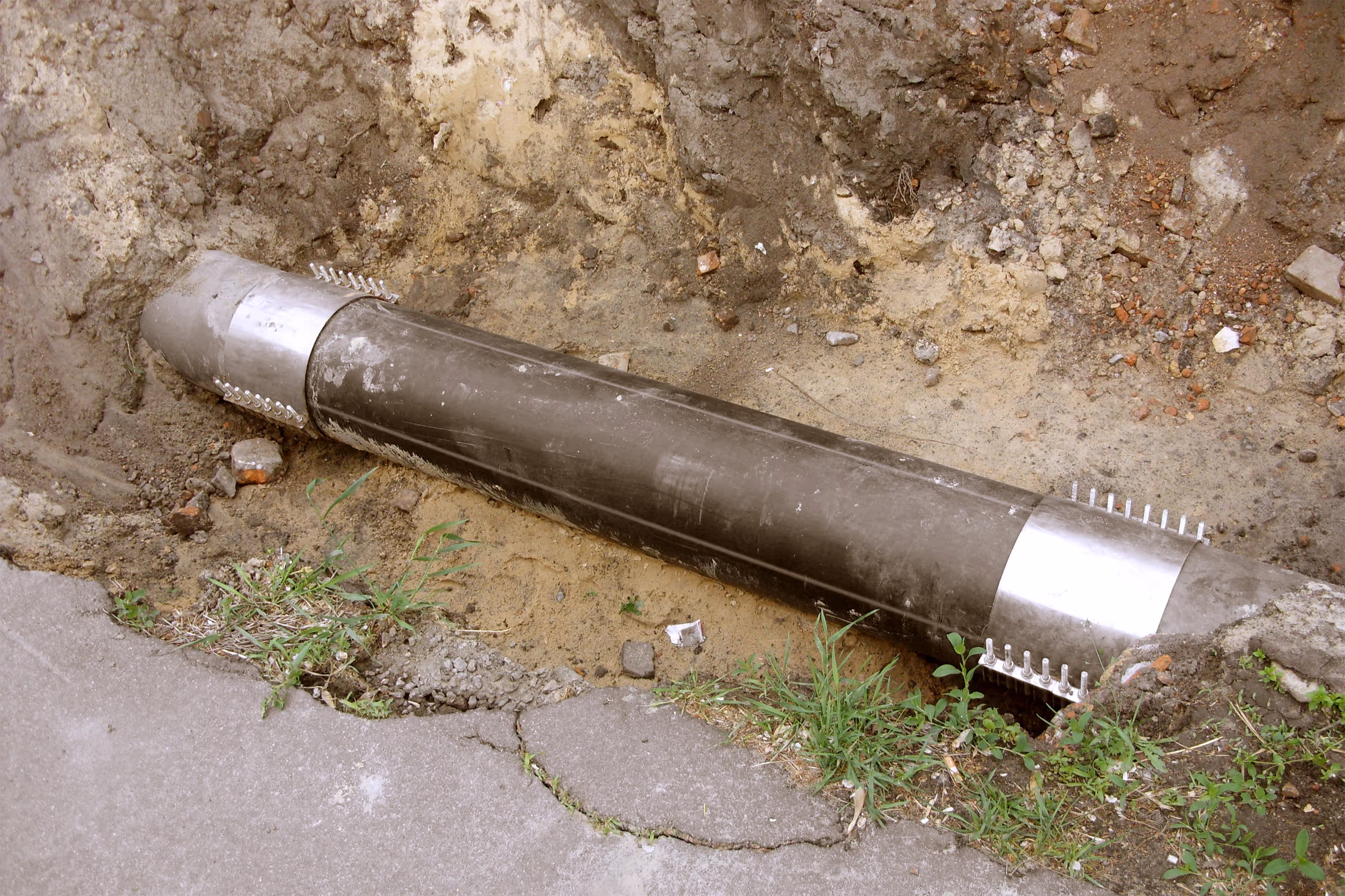 A large, dark metal pipe lies exposed in a dirt trench alongside a concrete pavement.