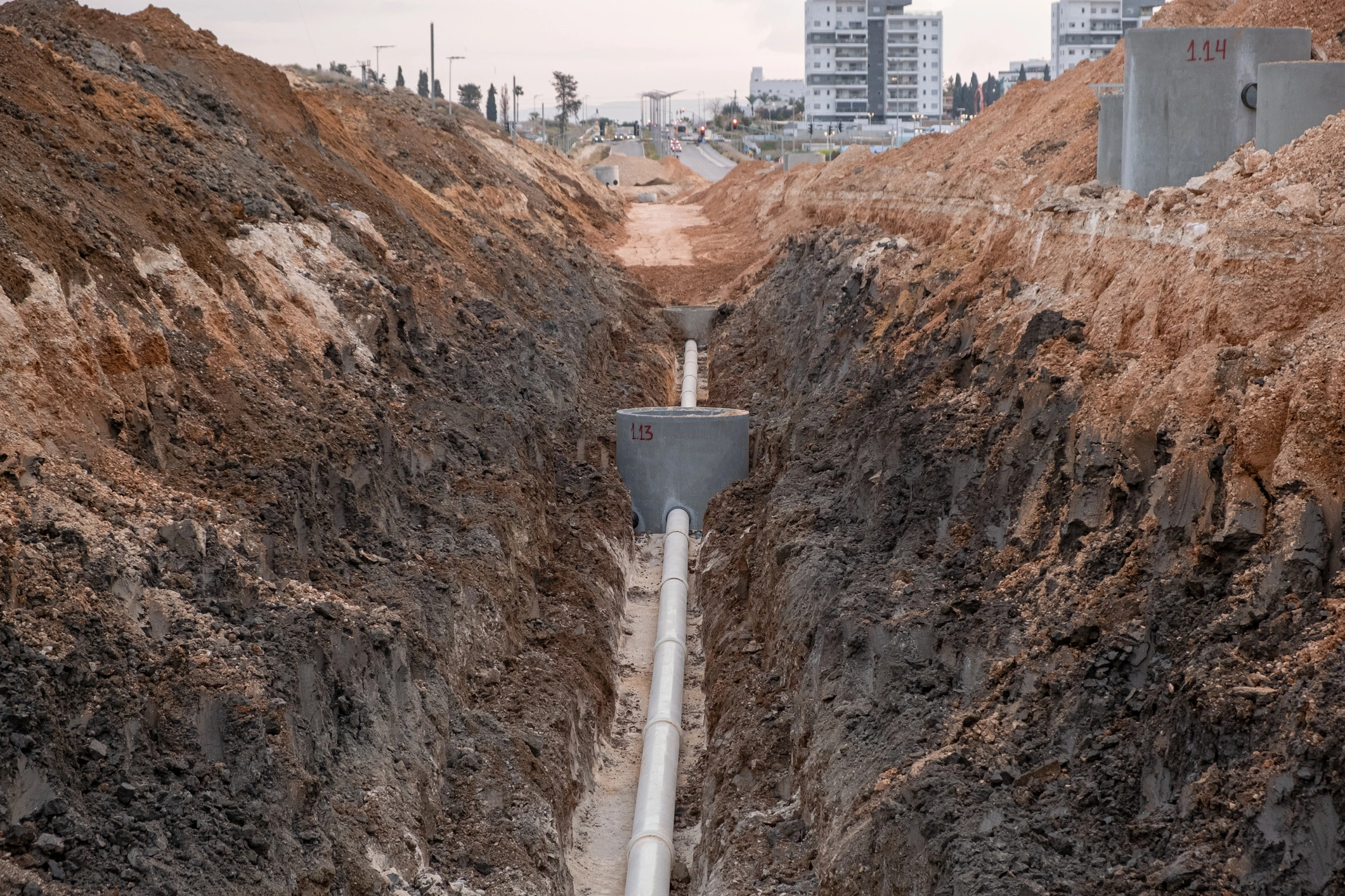 Trench with exposed pipeline stretches into distance, flanked by piles of earth, under an overcast sky.